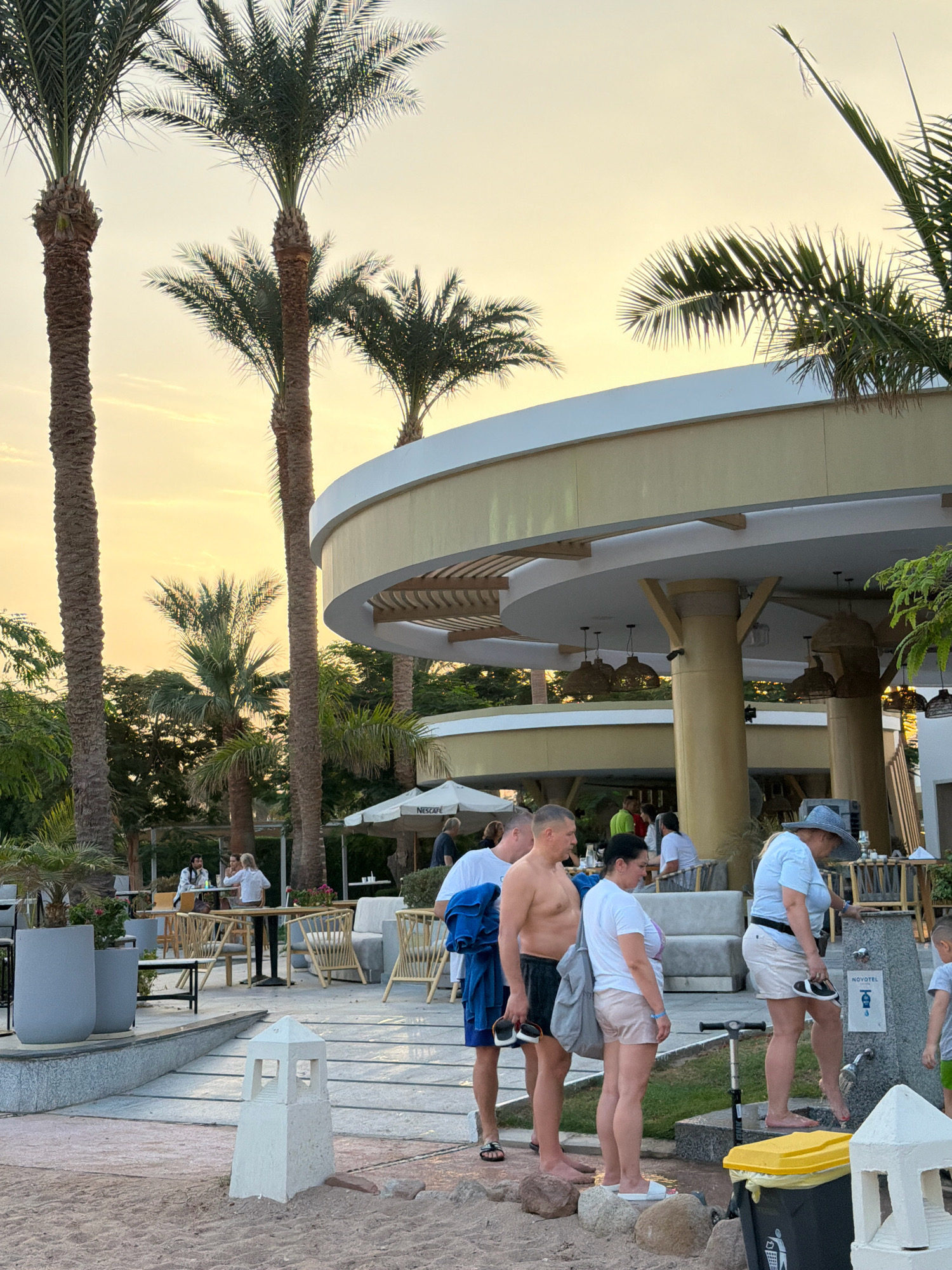 The image shows an outdoor area at a resort, likely in Sharm El Sheikh, Egypt, with palm trees and a partially visible building with an open-air dining or seating area. Several people are present. In the foreground, four individuals are standing near a water fountain. One man, bare-chested and wearing black shorts, holds a towel. A woman in a white t-shirt and pink shorts stands next to him, holding a grey bag. Another woman, wearing a light blue shirt and a straw hat, is using the water fountain, rinsing her feet. A young child in a grey t-shirt and green shorts stands nearby. In the background, several tables and chairs are visible under umbrellas and a covered structure. The sky is a light orange-yellow, suggesting it is either sunrise or sunset.