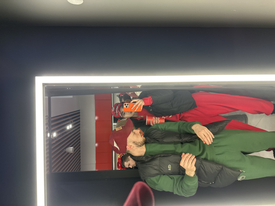 The image depicts a mirror selfie taken inside a retail store in Yekaterinburg, Russia, as evidenced by "URALMASH" branded caps worn by the subjects, referring to a Yekaterinburg sports club.
In the reflection, two individuals are prominent. On the left, a male subject with a short beard wears a maroon baseball cap with a light emblem, a dark green long-sleeved shirt, matching green sweatpants with a white logo, and a black puffer vest displaying a white Lacoste crocodile logo on the left chest. He looks downwards and to his right. On the right, a female subject takes the photograph using an orange-cased smartphone, obscuring most of her face. She wears a red baseball cap, a red zipped tracksuit, and a black puffer jacket. She holds a red disposable cup in her left hand.
The mirror is framed by vertical LED lighting. The reflected background reveals a store interior with red display shelving filled with merchandise (likely additional caps), a dark ceiling composed of slatted panels, and recessed lighting fixtures. A third male subject, partially visible behind the primary male, appears to wear a dark jacket and looks forward into the store space. The immediate area around the mirror inside the frame is dark-colored.