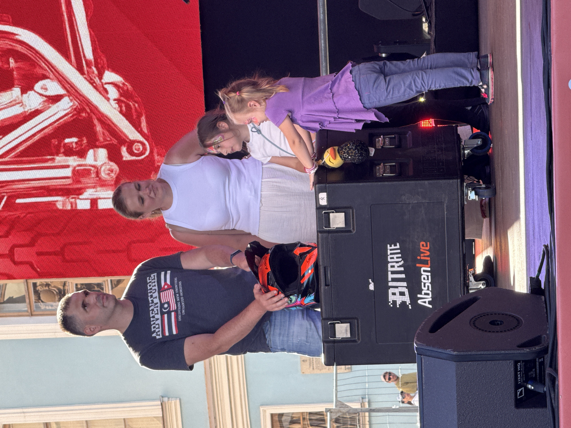 A family poses for a photo in front of a large red screen displaying a white graphic, possibly a vehicle. The scene appears to be outdoors, with a light blue building visible in the background and a street and fence. The time of day seems to be daytime, with bright lighting.
In the foreground, a man in a dark grey t-shirt with the text "ADVENTURE DISCOVERING NEW FRONTIERS UNCHARTED HORIZONS" stands to the left. He is holding an orange and black helmet. To his right, a woman in a white tank top is seen from the side. Two young girls are also present. One girl, in a purple top and blue jeans, is leaning over a black case with wheels. The other girl, wearing a white top, is looking down. A plush toy with a star pattern is on the black case.
The black case has the text "BITRATE AbsenLive" printed on its side, along with other smaller text and logos. Part of a black speaker is visible to the right of the case. The overall mood appears to be casual and happy, suggesting a family outing or event. The presence of the helmet and the "Adventure" text on the t-shirt might hint at an outdoor activity or event related to adventure sports.