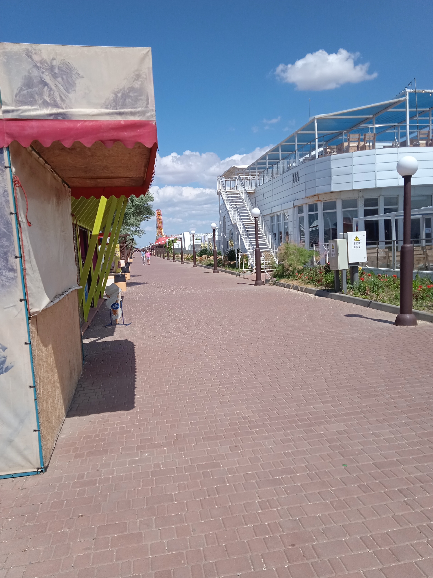This is a full-shot, outdoor, eye-level, daylight image of a paved promenade. On the left, a stall with a white awning and red trim is visible. The stall has a beige and light brown exterior with some vertical yellow supports. Beyond the stall, the promenade stretches into the distance, lined with lamp posts and vegetation on the right. A large white building with multiple levels and an open-air upper deck with blue covering is on the right side of the promenade. Several people are walking in the distance along the promenade. The sky is blue with scattered white clouds.