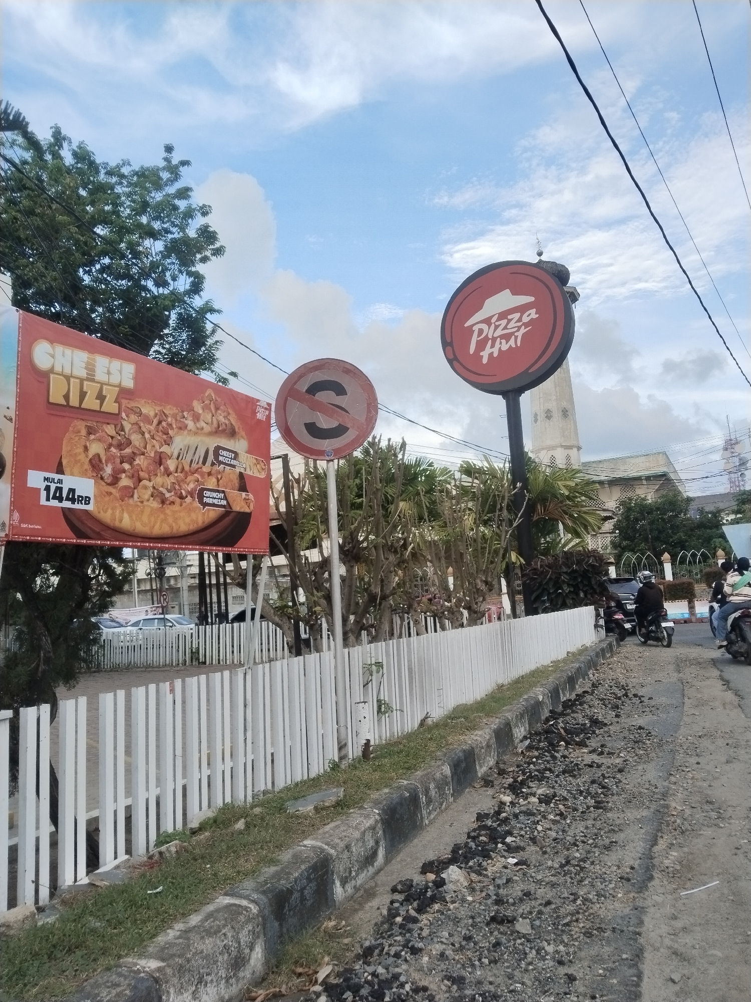 The image shows a Pizza Hut restaurant in Lhokseumawe, Indonesia. A "CHEESE RIZZ" advertisement is on display, as well as a "no stopping" sign. A white picket fence lines the sidewalk. Motorcyclists are traveling on a broken and uneven street. In the background is a tall, light-colored building with a tower. The sky is cloudy.