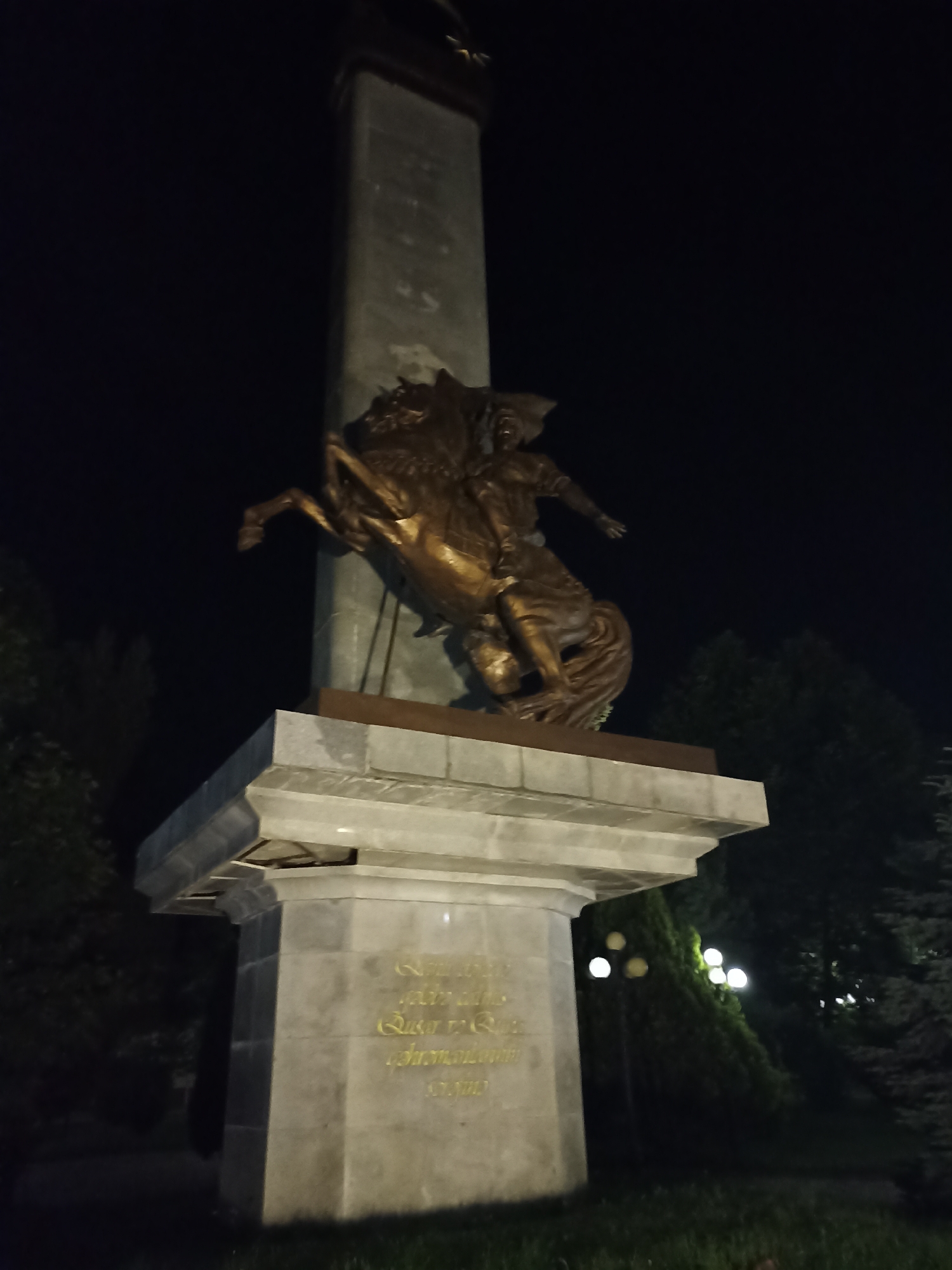 The image shows a nighttime view of the Monument to the Gusar Aviation Division in Gusar, Azerbaijan. The monument features a bronze sculpture of a soldier on horseback, with the horse rearing on its hind legs. The sculpture is positioned atop a tiered stone pedestal, with the main column of the monument rising behind it. The base of the monument has an inscription in yellow text, which is partially visible. The surrounding environment appears to be a park or public space, with trees and streetlights visible in the background. The lighting suggests the monument is illuminated for nighttime viewing.