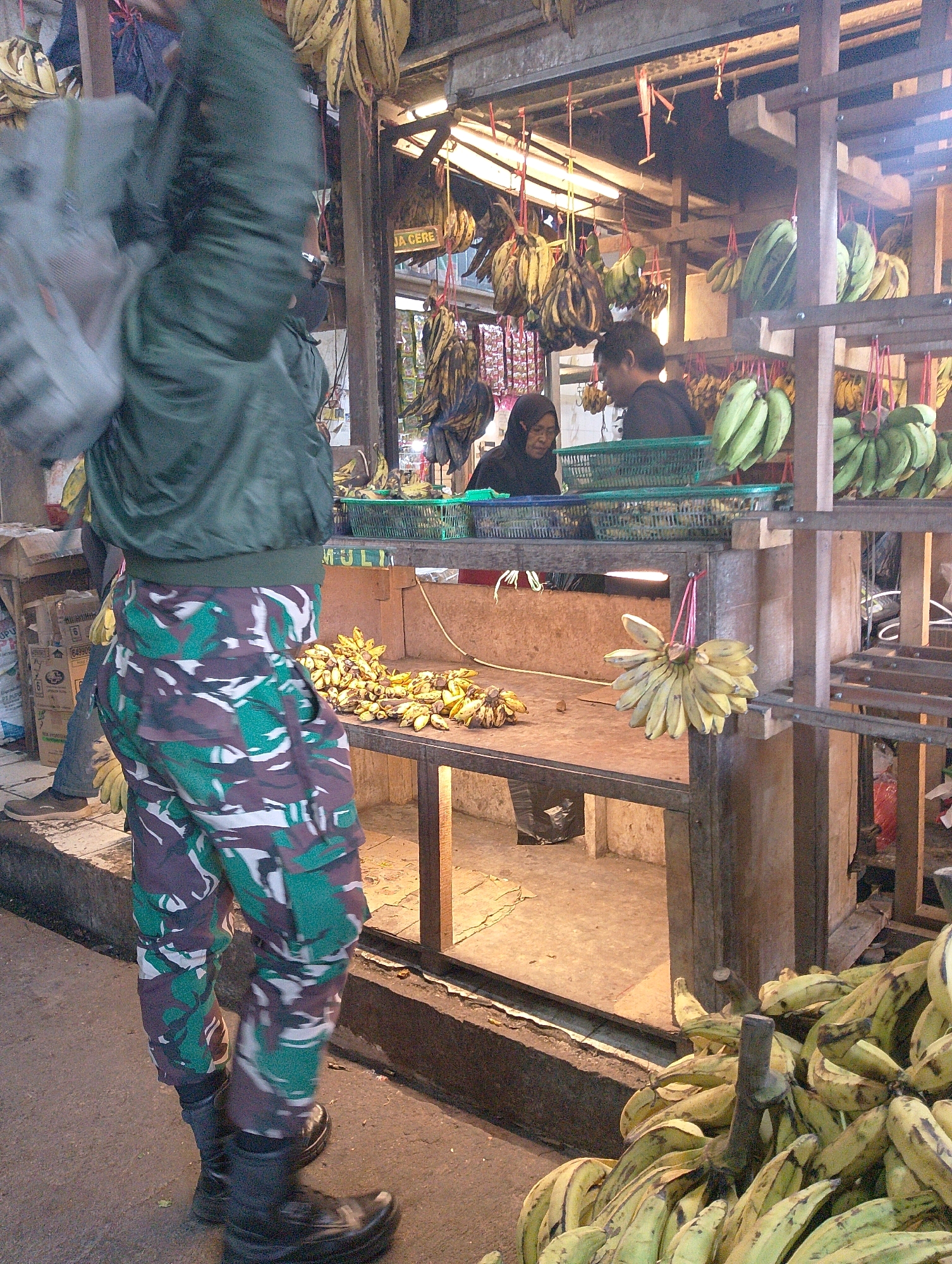 The image depicts an outdoor fruit stall in Lembang, Indonesia, likely a market. The foreground features a person wearing a camouflage patterned uniform, consisting of a green jacket and trousers with a mix of green, brown, and white camouflage. The person is walking away from the camera, their right arm raised, carrying a bag. The stall itself is constructed of wood and metal, displaying a large quantity of bananas. Clusters of yellow and green bananas are hanging from the ceiling and are piled on wooden tables. In the background, two individuals are visible behind the stall. One, wearing a black headscarf and dark clothing, appears to be a vendor. The other, a person with dark hair wearing a dark top, is partially obscured. The environment suggests a busy marketplace, with some boxes and other merchandise visible on the left side of the image. The lighting indicates it is daytime.
