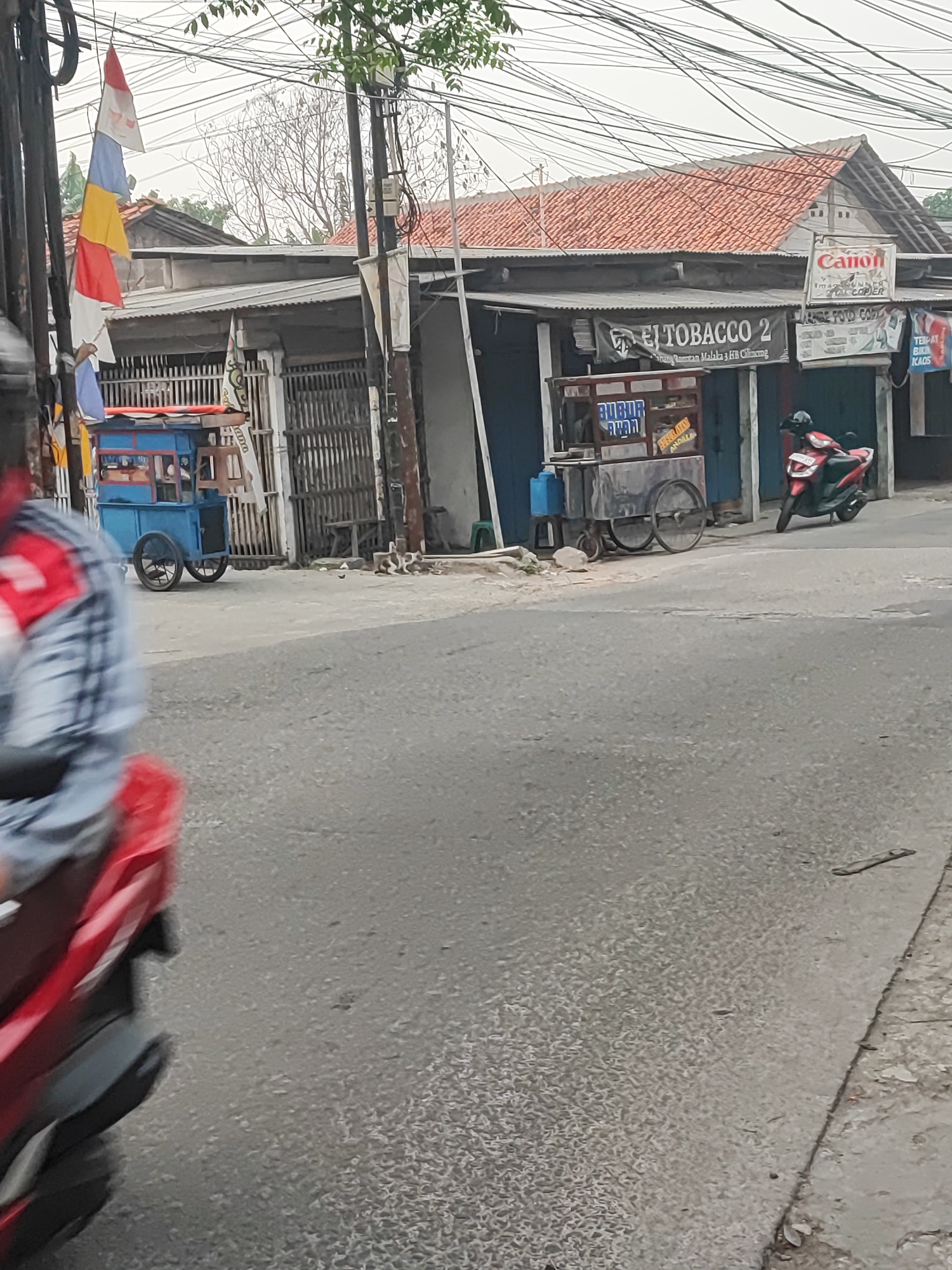 In Satriamekar, Indonesia, a street scene unfolds with a focus on a row of small storefronts and a street. In the foreground, a red motorcycle is partially visible, with its rider in a blurred motion, suggesting movement. To the left, a blue food cart with red trim is parked on the side of the road, its wheels visible. Above and behind the food cart, a series of Indonesian flags, predominantly red and white, are strung on a line. Further down the street, a red and white sign with the word "Canon" is displayed on one of the buildings, alongside text indicating "IMAGE PRINTER PHOTO COPIER." Another sign reads "BUBUR AYAM" which translates to "chicken porridge," likely indicating the type of food sold at a nearby cart or establishment. A dark blue awning extends over one of the shops. A red-tiled roof is visible on a building in the background. The street is paved and appears to be in moderate condition, with some debris present. Overhead, a dense network of electrical wires crisscrosses the sky.