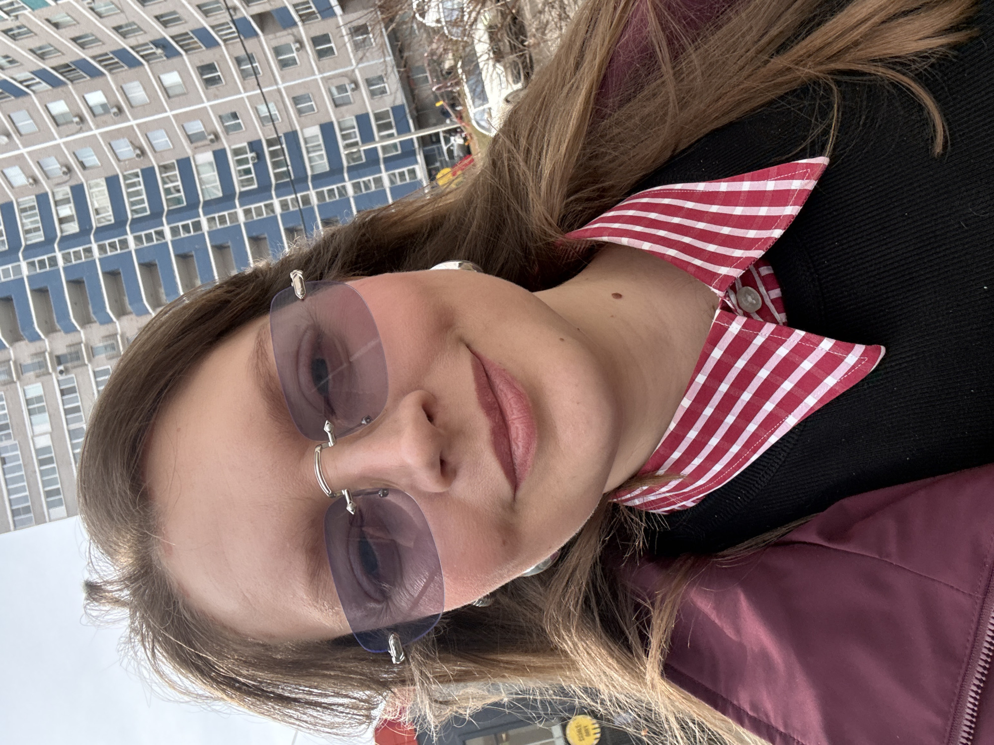 A medium close-up photograph shows a woman wearing rimless purple sunglasses, a black sweater over a red and white gingham collared shirt, and a plum-colored jacket. She is slightly smiling. The background reveals an urban setting, featuring a large residential building with numerous windows. This building resembles a typical Soviet-era apartment block. The location is identified as the city of Izhevsk, Russia.