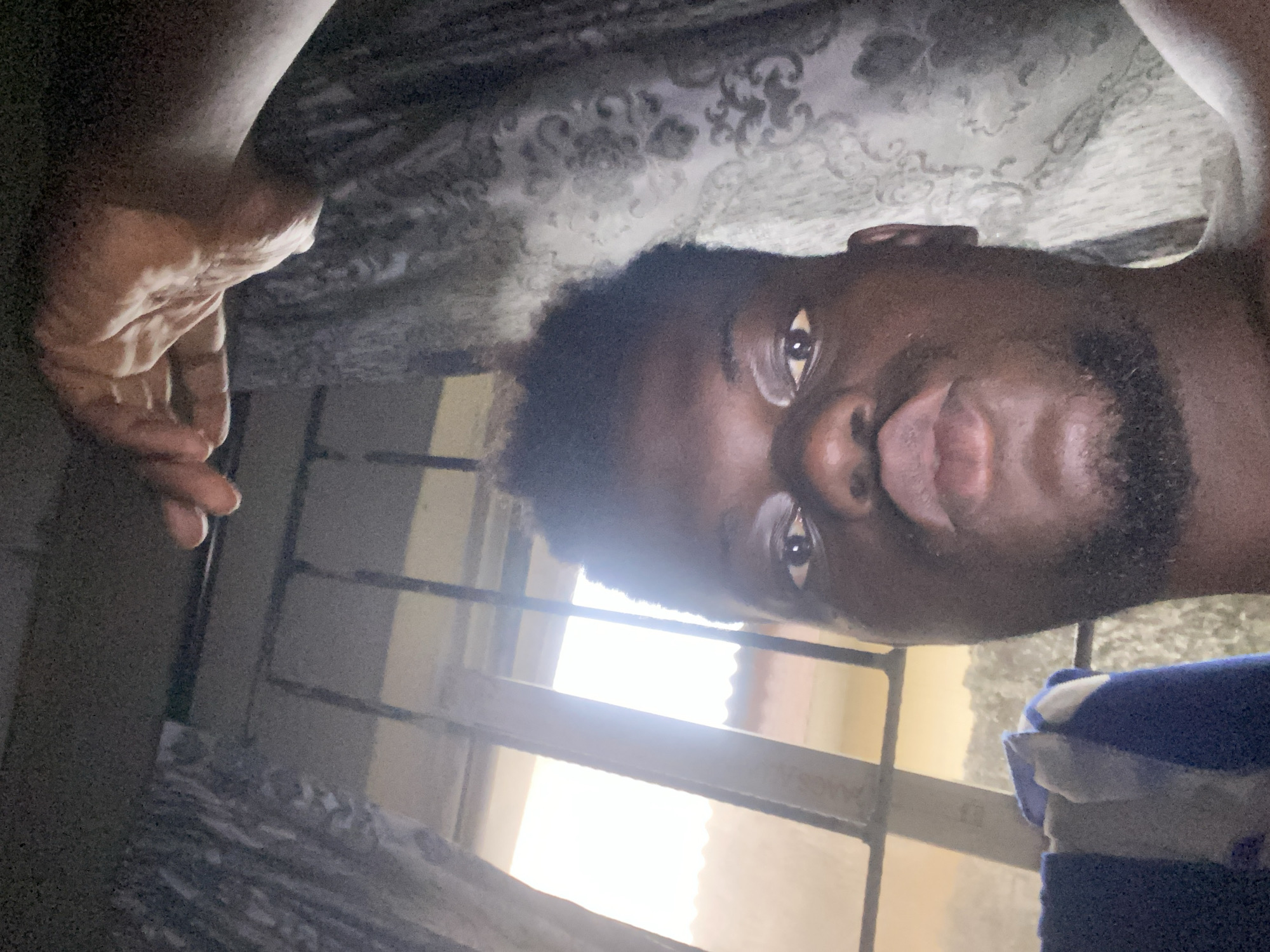 A selfie of a dark-skinned man with short, dark hair and a beard is angled upwards, capturing his face and a portion of his torso. His hand is partially visible on the left. He appears to be indoors, with a window behind him letting in bright light. Part of a blue and white striped garment is visible on the right, and patterned fabric can be seen above his head. The location is Unknown, Unknown.