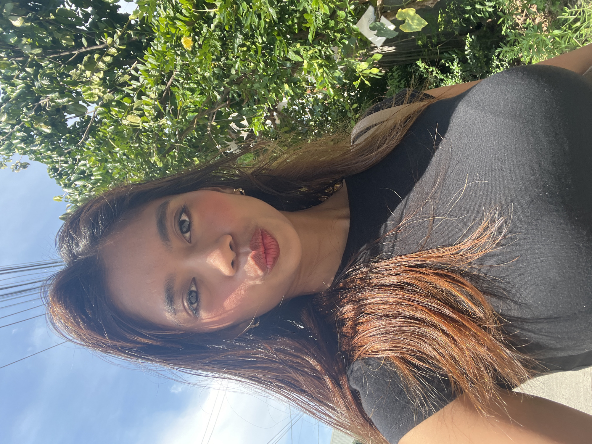 A young woman is the central subject of this close-up outdoor selfie, taken on a bright, sunny day in what appears to be Davao City, Philippines. She has long, dark brown hair with prominent reddish-brown highlights, flowing around her shoulders and chest, and is wearing a black top. Her face is angled slightly upwards, with her lips pursed in a playful "kiss face" expression. Her eyes are light-colored, possibly green or hazel, and she has visible makeup, including eyeliner and lipstick. A small, golden earring is visible in her left ear.
The scene is intensely lit by direct sunlight, casting strong highlights on her hair and skin, and creating distinct shadows across her face and body. Behind her, a dense expanse of lush green foliage from trees or bushes occupies the upper and right side of the frame, suggesting a tropical environment. To the left and below, a clear, bright blue sky with faint clouds is visible, crisscrossed by several dark utility lines.
Among the background foliage, a small, white, rectangular label is partially visible, containing faint, unidentifiable text (e.g., "GU" or "0U") and a simple drawing, possibly of a leaf. The overall impression is one of a cheerful, casual moment captured outdoors under strong daylight.