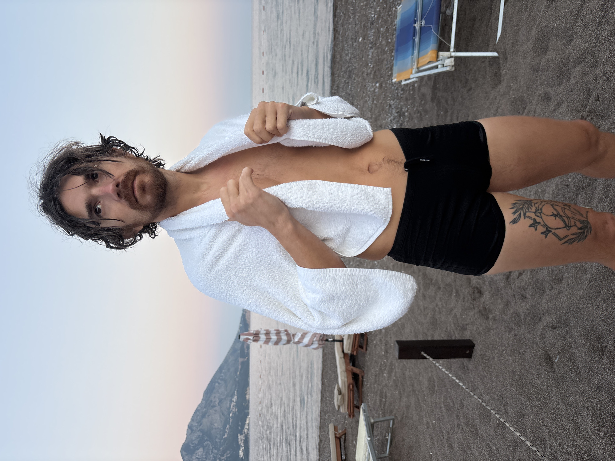 A man with wet, dark hair and a beard stands on a dark sand beach, wrapped in a white towel. He is wearing black swim trunks. His expression is neutral, and he is looking directly at the camera. The scene is set by the sea, with calm water visible in the background. Beyond the water, a mountainous coastline can be seen, suggesting a Mediterranean or similar climate. Several beach chairs and umbrellas are scattered on the sand, indicating a public beach area. The sky is overcast with soft, diffused light, possibly suggesting dawn or dusk. A tattoo is visible on the man's left thigh. There are no visible signs or text in the image. The overall mood is relaxed and casual.