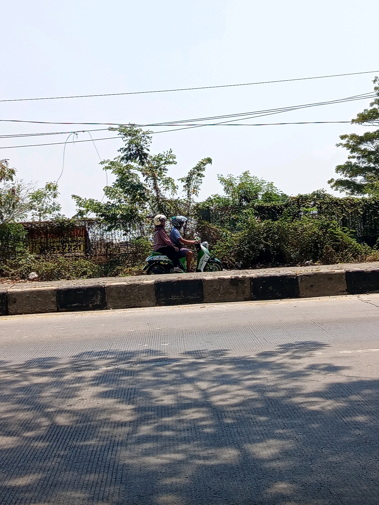Two individuals wearing helmets are riding a green and white scooter on a paved road. The person in the back is wearing a maroon shirt and the person in the front is wearing a blue shirt. They are traveling from left to right. The road is bordered by a concrete barrier with black and gray rectangular blocks. Behind the barrier, there is dense green foliage, some of which is growing over a rusty metal fence. Overhead, power lines are visible against a bright, hazy sky. The foreground of the image shows the road surface with a pattern of textured lines and dappled shadows cast by trees. This scene is consistent with a location in Karawang, Indonesia, likely along a roadside with vegetation.