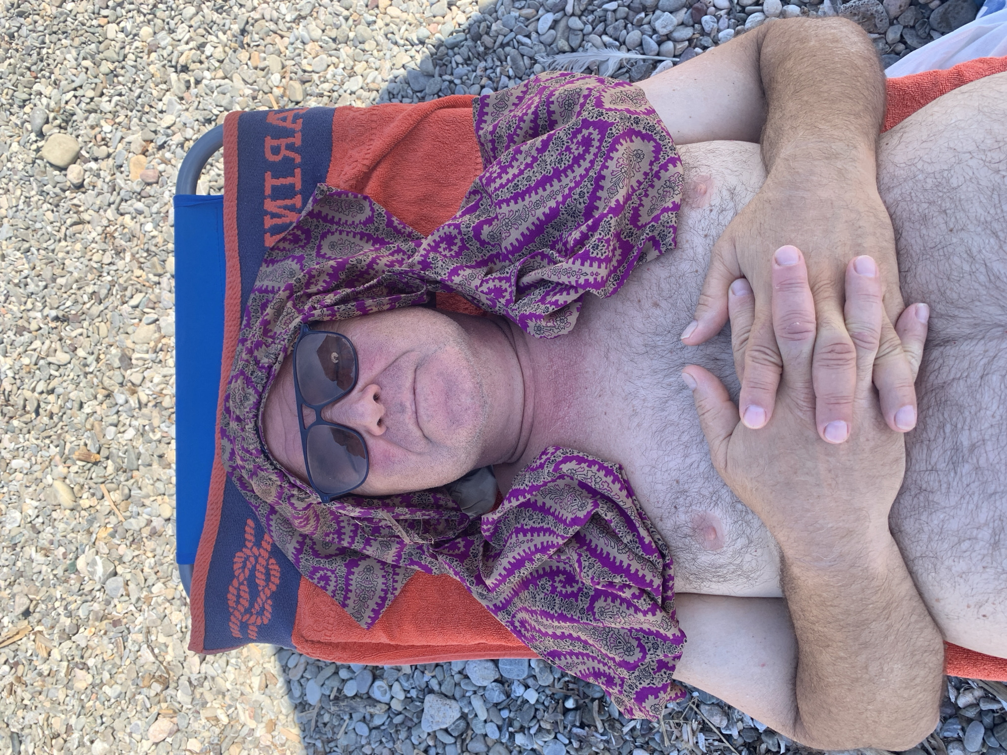 The image shows a person lying on a beach chair, covered by an orange towel and a patterned cloth. The person is wearing sunglasses and their arms are crossed over their chest. The scene is set on a beach with many small pebbles. The time of day appears to be sunny, as suggested by the sunglasses and the bright lighting. The location context is Pozzarello, Italy, which is consistent with a beach setting. No specific emotions are overtly displayed, beyond a relaxed pose. There is visible text on the blue part of the towel, which appears to spell "MARTINI" when read from right to left, with some letters obscured.