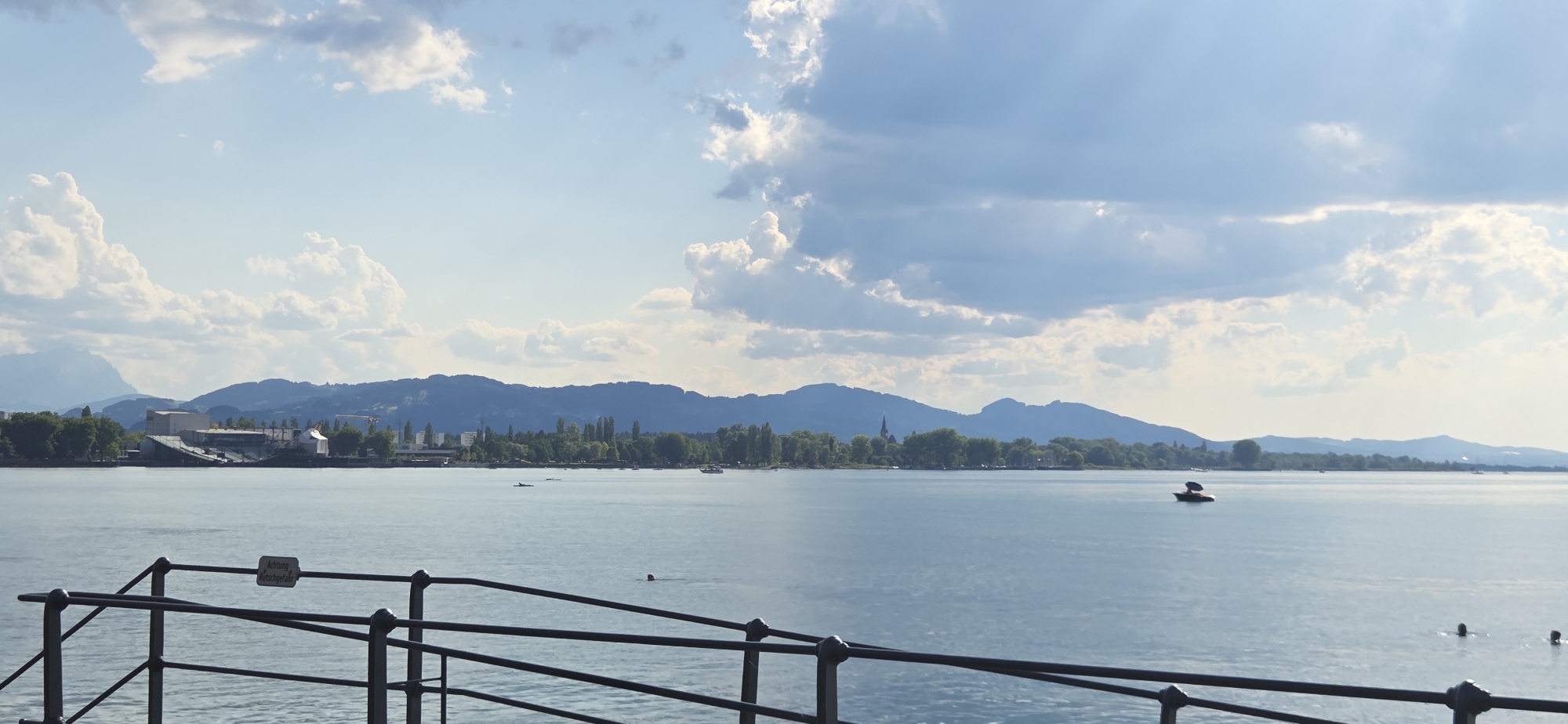 This media file captures a serene daytime scene on a large body of water, likely a lake, with a distant shoreline featuring a town, lush greenery, and rolling hills or mountains. The sky is filled with a mix of white, fluffy clouds and some darker, more dramatic clouds, suggesting a dynamic weather pattern. The sun is out, casting a gentle light on the water, which appears calm and reflects the sky.
In the foreground, a dark metal railing with circular posts creates a barrier, suggesting a pier or a walkway. A sign is attached to the railing, partially visible, with German text that reads "Achtung Rutschgefahr," meaning "Caution, Risk of Slipping."
On the water, several objects and subjects are visible. A boat with a dark canopy is sailing in the middle distance. Further back, smaller boats are scattered across the water. There are also individuals swimming or wading in the water, indicated by dark shapes of heads or bodies just above the surface.
The shoreline is a mix of natural and man-made elements. Dense green trees line the water's edge, interspersed with buildings that appear to be part of a town or industrial area. The distant backdrop is dominated by hazy blue mountains, contributing to the sense of depth and scale of the landscape. The overall atmosphere is peaceful and inviting, with a hint of activity from the boats and swimmers. The time of day appears to be late afternoon or early evening, given the soft lighting and the presence of clouds. The location cues, such as the German text and the mountainous landscape, suggest a Central European setting, consistent with the provided context of Lochau, Austria.