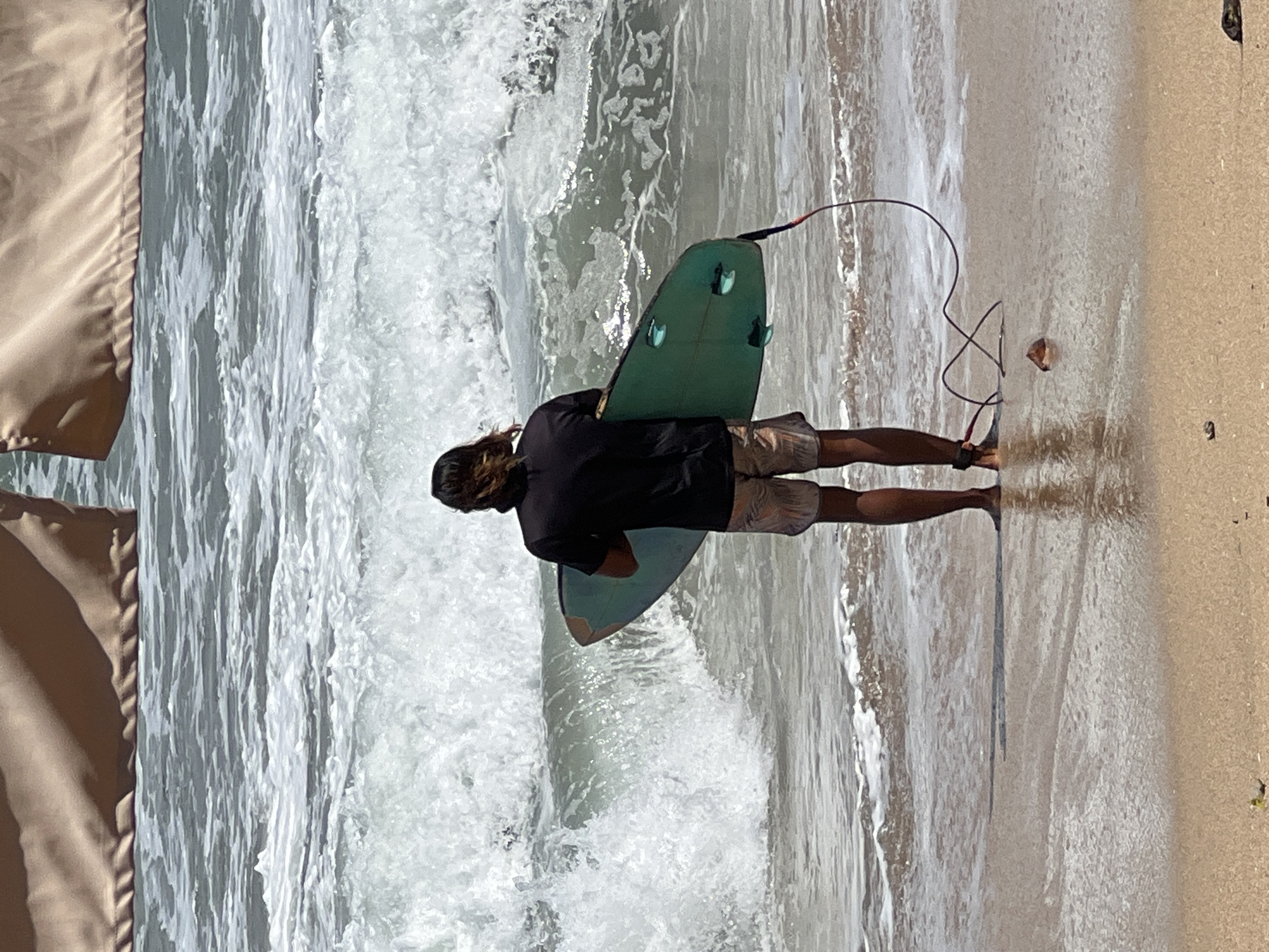 The image shows a lone surfer standing on the shore of Kuta Beach, Indonesia, holding a teal surfboard. The surfer is facing away from the camera, towards the ocean, with the surfboard tucked under their right arm. They are wearing a black t-shirt and patterned swim trunks. The ocean is active with crashing waves and white foam. The sand is wet at the waterline, reflecting the light. In the background, a tan fabric structure, possibly part of a beach umbrella or canopy, is visible at the top of the frame. A dark leash trails from the surfboard to the surfer's ankle. The overall scene captures a moment of anticipation before entering the water.