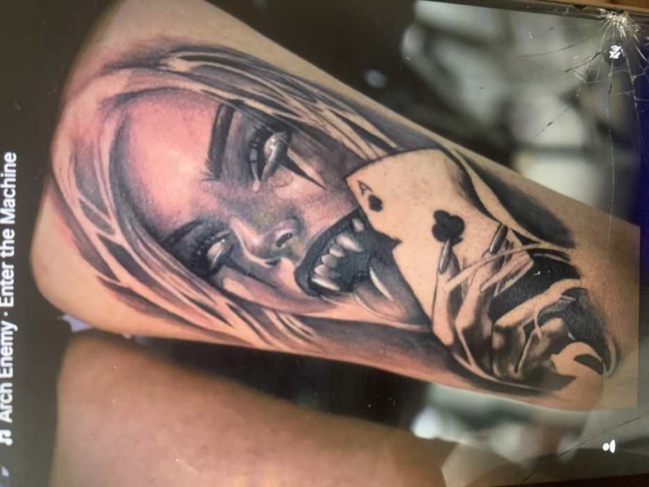 The image displays a photograph of a monochrome tattoo with subtle color accents on a person's skin, likely an arm or leg. The tattoo depicts a stylized female face with long, flowing hair, wide-open eyes featuring bright white irises and pupils, and a mouth open to reveal sharp fangs and an extended tongue. Dark, lightning bolt-like marks are visible below the outer corners of her eyes. Her skin in the tattoo appears pale, with subtle reddish-purple shading around the hairline and cheeks. A hand with long, dark nails, belonging to the depicted figure, holds an Ace of Clubs playing card. The top edge of the card appears to be held between the figure's fangs.
The photograph itself appears to have been taken of a digital screen, indicated by text "Arch Enemy - Enter the Machine" visible at the top edge and a prominent crack in the bottom-right corner of the screen. The skin surrounding the tattoo shows minor redness, consistent with a recently applied tattoo. The image does not contain any visual or textual information to determine its specific geographic location, including the city of Goliam Izvor, Bulgaria.