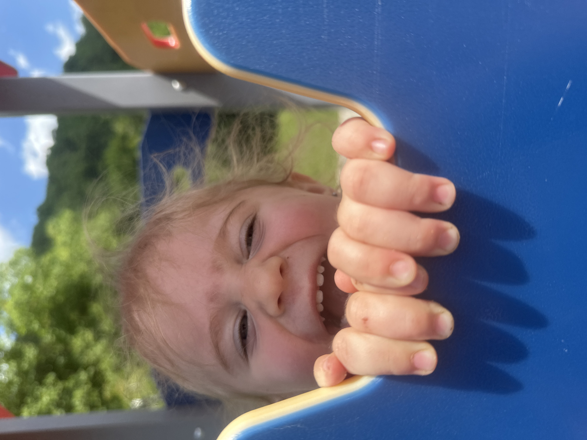 A close-up, low-angle shot captures a toddler with blonde hair leaning into a blue and yellow playground structure, their hands gripping the edge. The child's face is partially obscured but shows a wide, open-mouthed expression with visible teeth and rosy cheeks, eyes squinting. The background is blurred, revealing green foliage and a bright blue sky with white clouds, suggesting an outdoor setting on a sunny day. The specific location in Donchovci, Bulgaria, cannot be determined from the visual information provided.