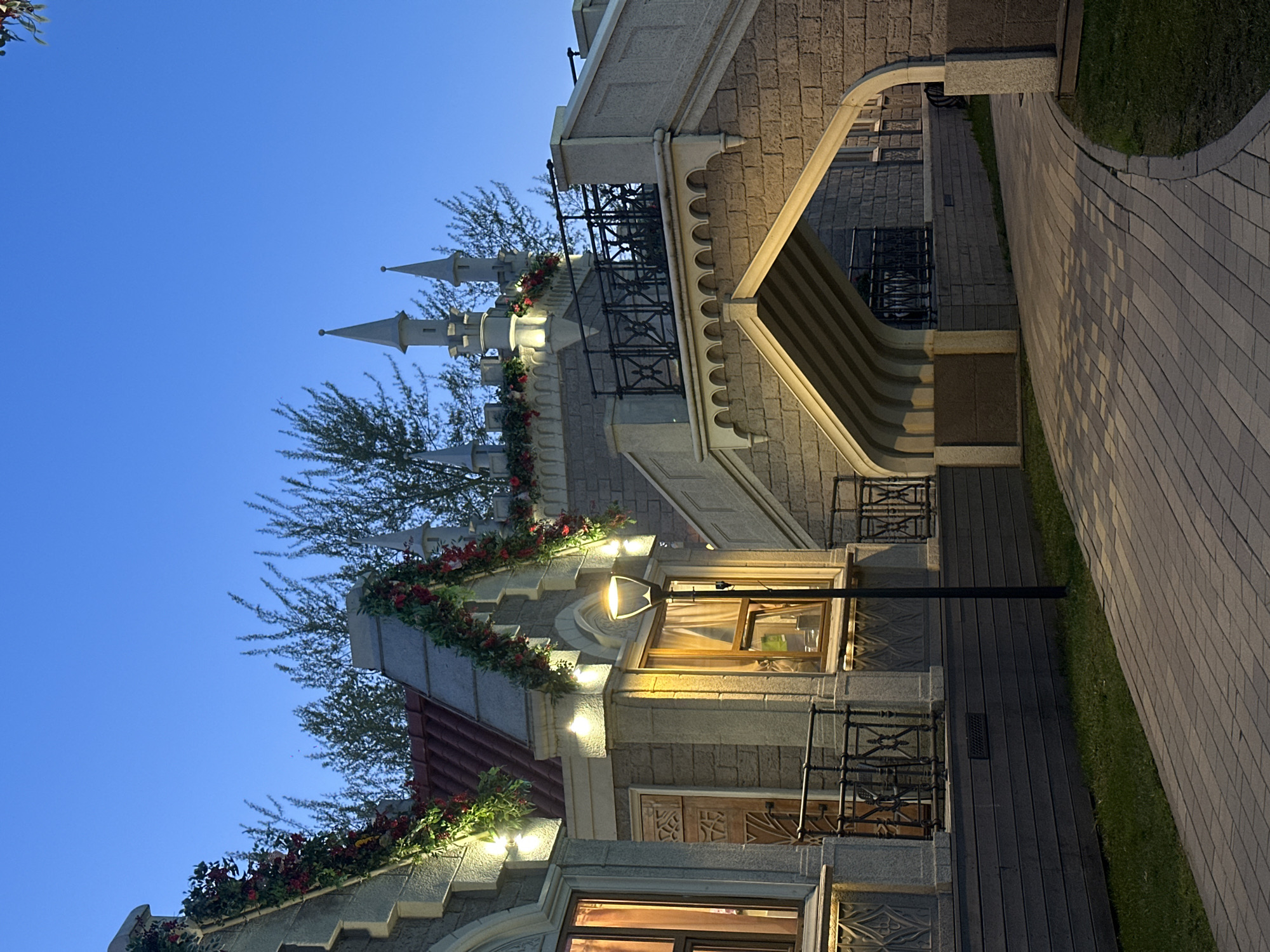 A nighttime exterior shot shows architectural details and landscaping in Moscow, Russia. The architecture, composed of brick and stone, is stylized with turrets reminiscent of a castle, decorated with greenery and garlands. A lamppost illuminates a window, and other windows are also lit. Railings and a bridge-like structure are visible. The sky is a deep blue, suggesting twilight. The landscaping includes grass and paved walkways.
