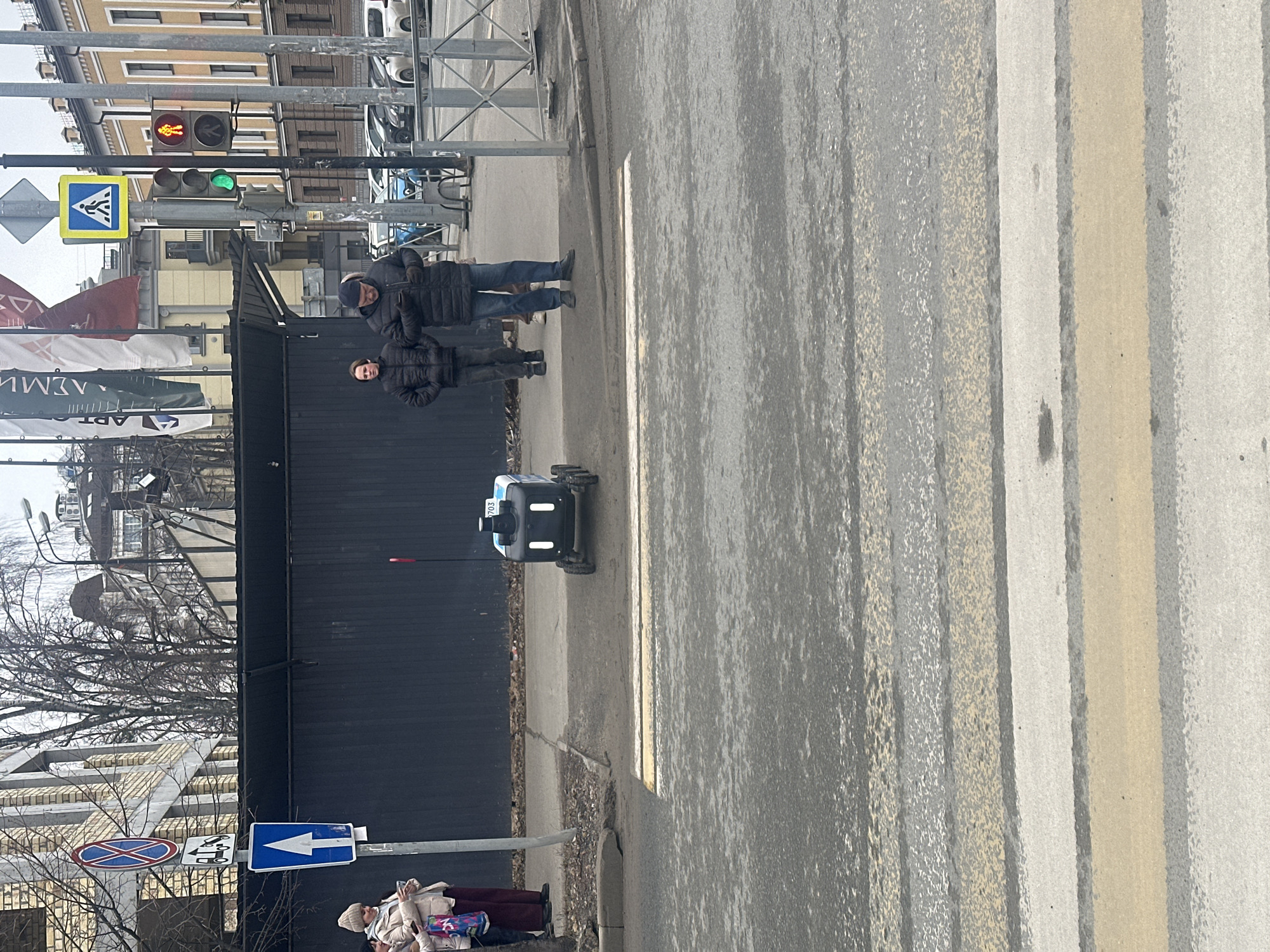 A street scene in Kazan, Russia shows an intersection with a crosswalk. Two men and a boy stand on the sidewalk next to a black fence. A delivery robot with white and blue accents is positioned on the sidewalk. A traffic light shows green for pedestrian crossing. A pedestrian crossing sign is also visible. At the lower left of the frame, an elderly woman is standing on the sidewalk. A blue and white street sign is nearby. In the background, there are buildings with yellow facades and leafless trees. The crosswalk is marked with alternating white, grey and tan stripes.