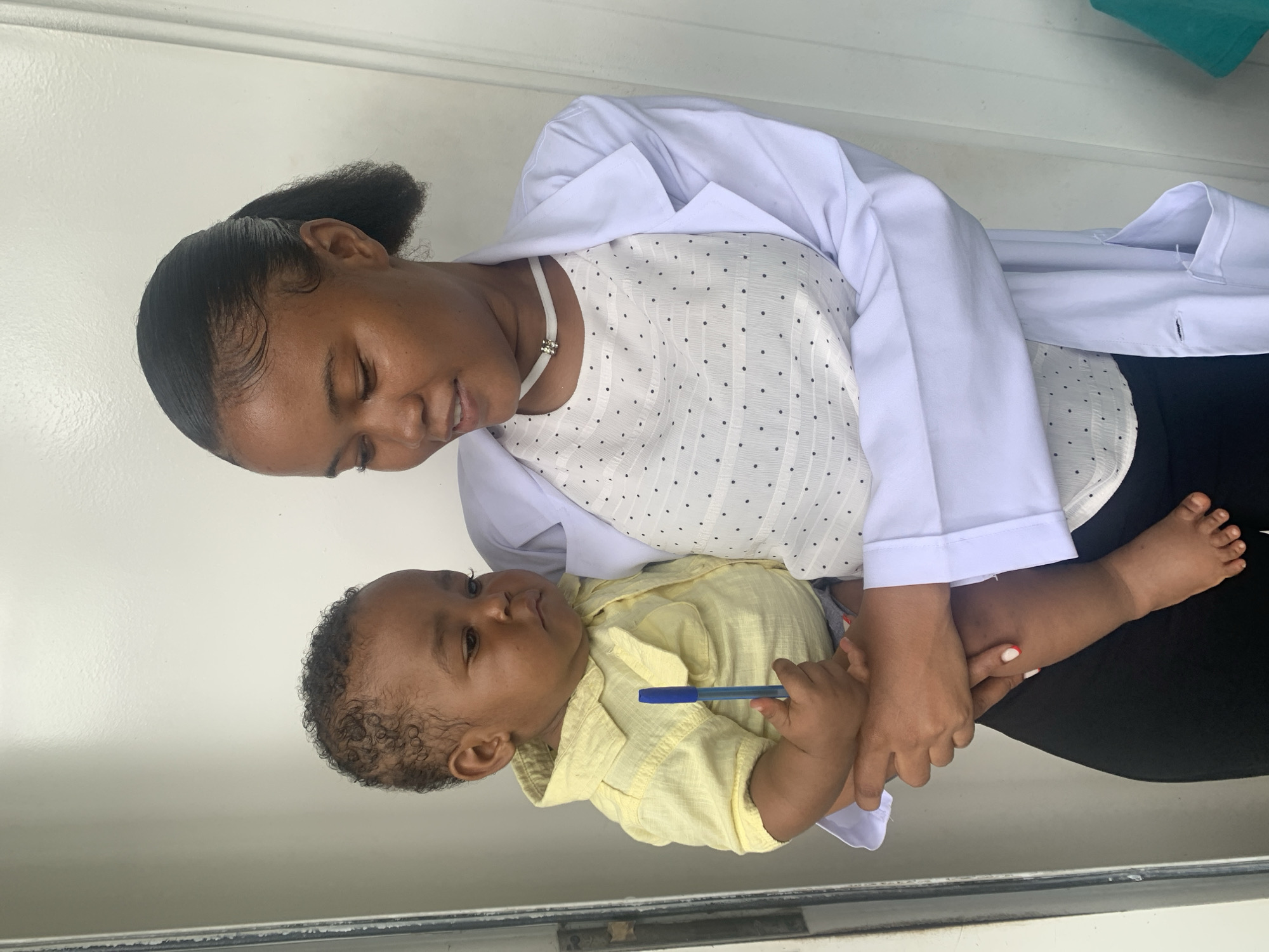 The image shows an adult female holding an infant against a plain, light-colored background. The adult, a young woman with dark skin and dark hair styled in a sleek ponytail, wears a white lab coat over a white, short-sleeved blouse with black polka dots, a white choker necklace, and black trousers. She looks downward towards the infant, with a slight smile. Her right hand, visible with light-colored nail polish, supports the baby's back. The infant, a baby boy with dark skin and curly hair, wears a light yellow collared shirt and gray shorts. He holds a blue ballpoint pen in his right hand and looks slightly to his right with a neutral expression. The background consists of a featureless light-colored wall or door, with a dark vertical line indicating a frame or edge on the left. No specific visual identifiers within the image indicate its location in Dar es-Salaam, Tanzania.