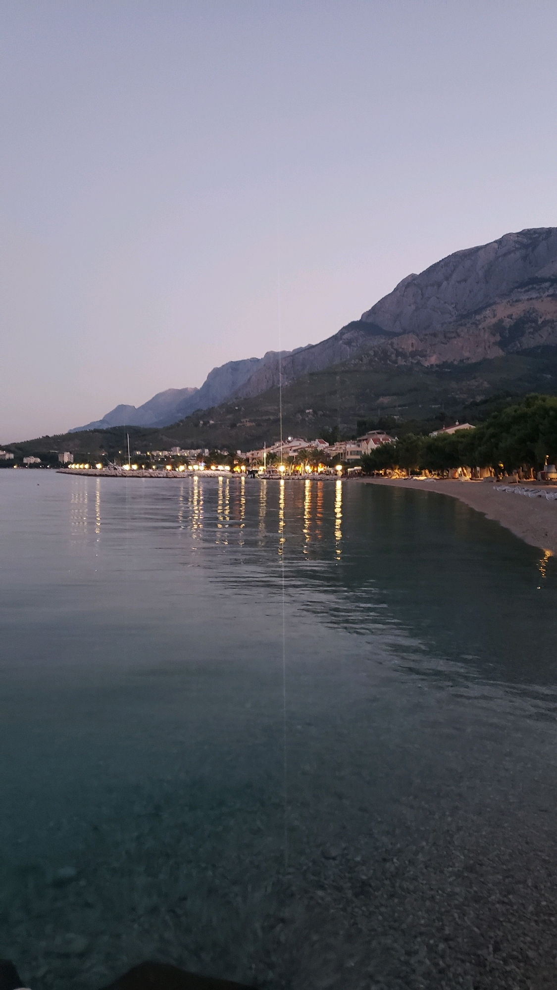 The image depicts a tranquil evening scene at a coastal town in Općina Tučepi, Croatia. The sky is a soft gradient of pale blue and purple, indicative of twilight. Majestic, rugged mountains rise in the background, their dark silhouettes contrasting with the lighter sky. Along the base of the mountains, a string of buildings with illuminated windows and lights line the shore, suggesting a developed settlement. A beach with rows of lounge chairs is visible in front of the buildings. The foreground is dominated by the calm, clear water of the Adriatic Sea, with the pebbled seabed faintly visible beneath the surface. Reflections of the town lights shimmer on the water's surface, creating a mesmerizing visual effect. The scene conveys a sense of peace and serenity, likely captured during the early evening hours after sunset.