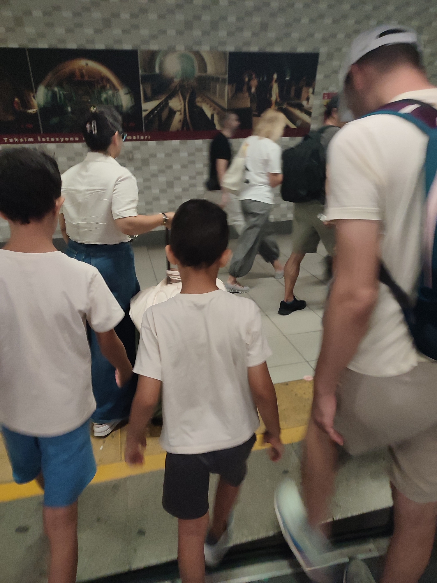 In Beyoğlu, Turkey, two young boys wearing white t-shirts and shorts, one blue and one dark, walk away from the camera down a subway platform. Behind them, a woman in a white top and dark pants, and a man in a light-colored t-shirt and light-colored shorts with a backpack, also walk away. The background features a tiled wall and posters depicting subway tunnels and stations.