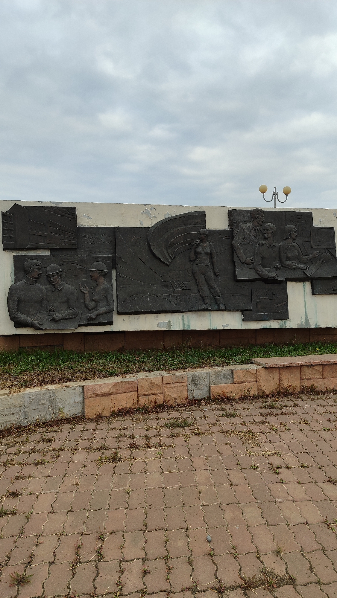 This is a vertical, medium-shot, eye-level photograph of a mural on a concrete wall in Mirny, Russia, on an overcast day. The mural is a bas-relief sculpture depicting figures in a stylized, socialist realist manner. The mural is divided into several panels. On the far left, there is a panel depicting two men and a woman in work clothes, appearing to be in discussion. To the right of this, a larger panel shows a woman in athletic attire, with a flag or banner unfurling behind her. To the right of that, two smaller panels show groups of people, one with a man and a woman, and another with a man and two women, all appearing to be in conversation or engaged in an activity. Above the mural, a lamppost with two yellow globes is visible. The mural is mounted on a concrete wall that shows signs of wear and peeling paint. Below the mural, there is a strip of grass and a decorative border made of rough-hewn stone blocks. In the foreground, there is a paved area with interlocking, brick-red pavers, with sparse weeds growing between them. The sky is filled with gray, textured clouds.