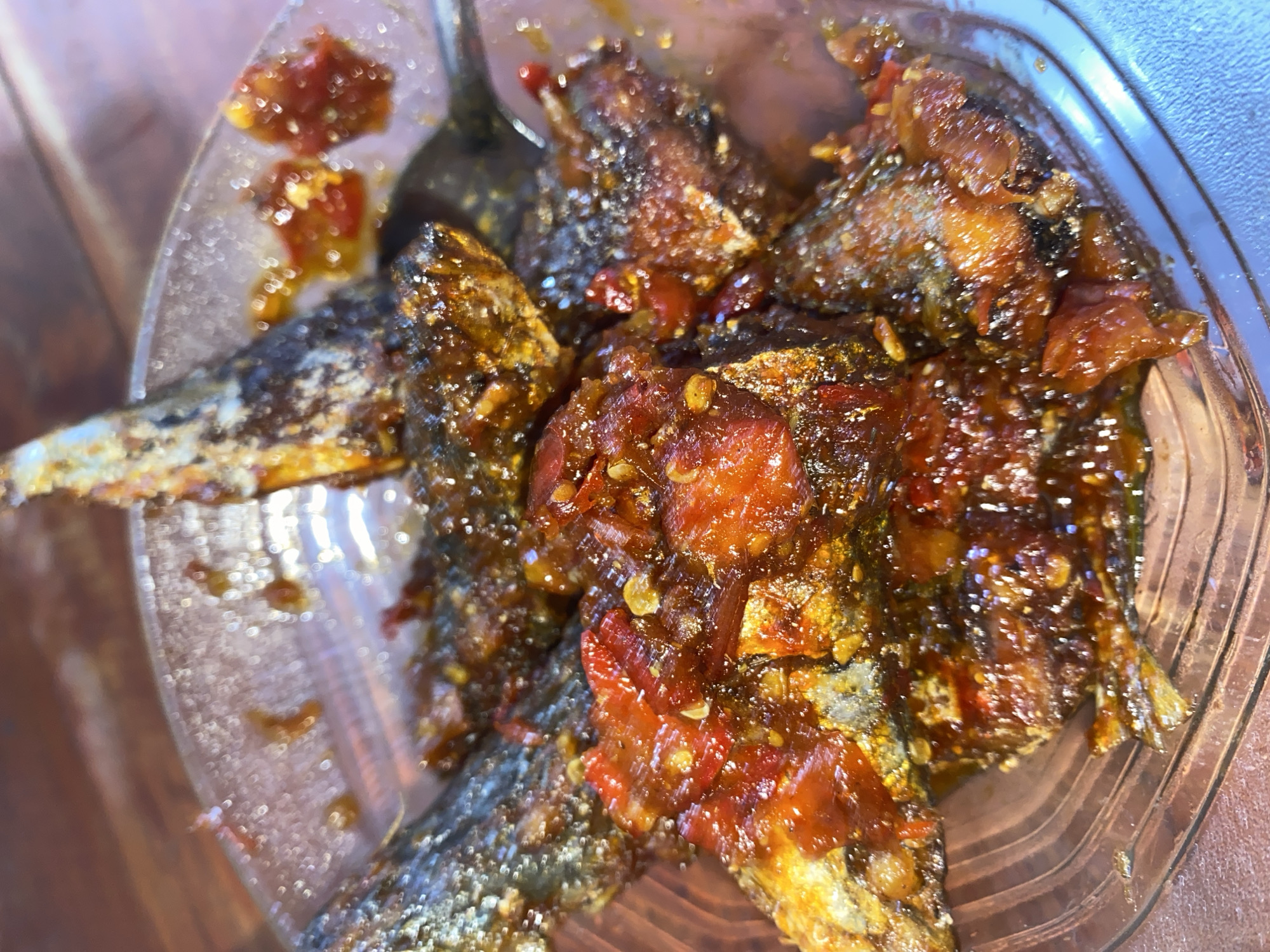 A close-up, overhead shot shows a clear, round glass bowl filled with several pieces of fried fish coated in a reddish-orange sauce. The fish appear to be small, whole fish, with visible fins and tails. The sauce is thick and glossy, with chunks of what appear to be chili peppers and other spices mixed in. A silver fork is partially visible in the upper right of the bowl, resting on the fish. The bowl is sitting on a wooden surface, likely a table, which has a textured grain. The lighting is warm, casting highlights on the glossy sauce and the smooth surface of the bowl. The background is slightly blurred, but the wooden texture is still discernible. This image was captured in Ta', Indonesia.