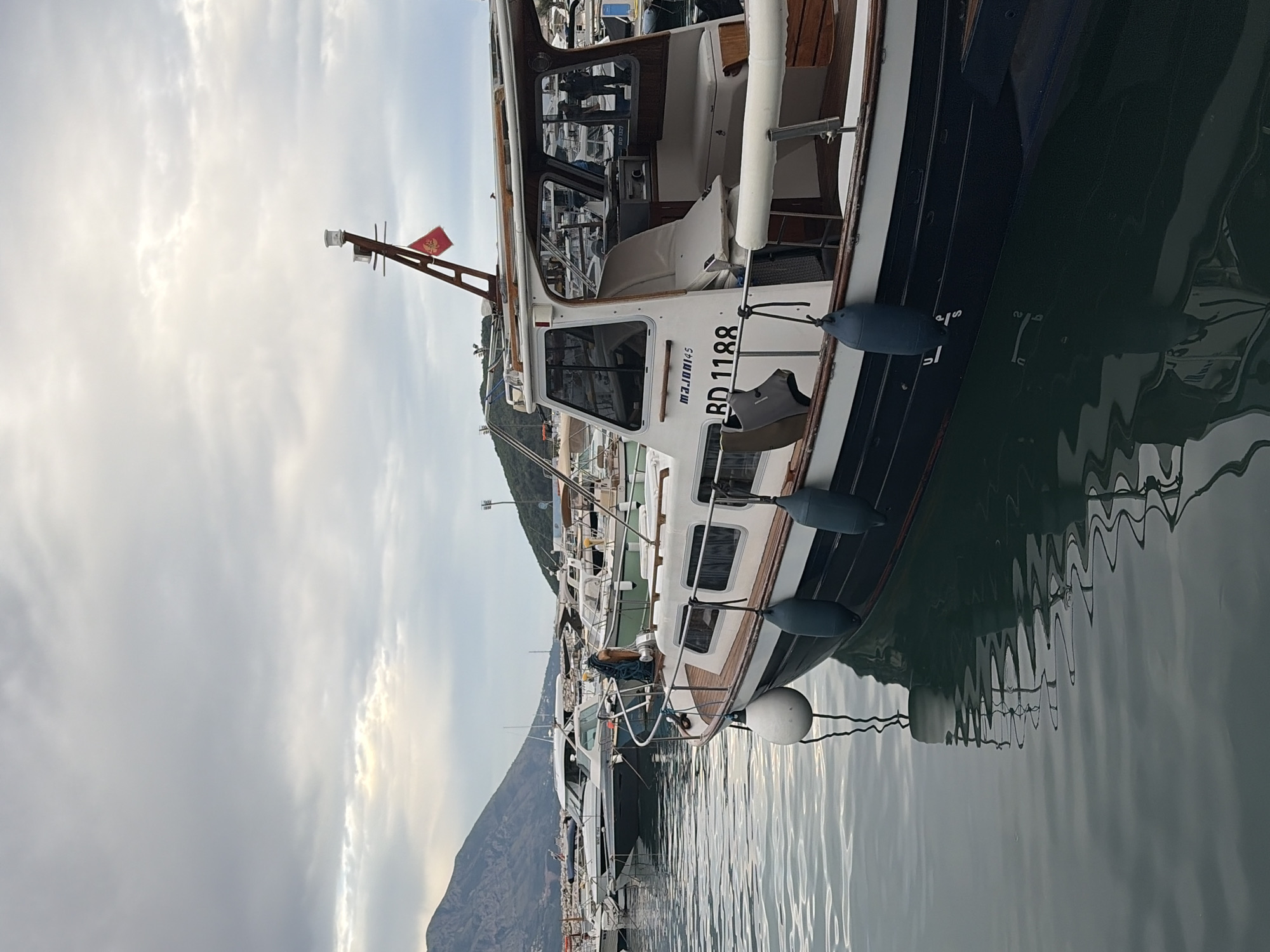 The image shows a marina filled with yachts and boats, likely located in the city of Budva, Montenegro, given the coastal setting and mountainous backdrop. The sky is overcast with a mix of gray and white clouds. Several boats are moored side-by-side, with the foreground boat showing details such as hull colors, fenders, windows, and a flag. Water reflects the boats and sky, creating a rippled effect.