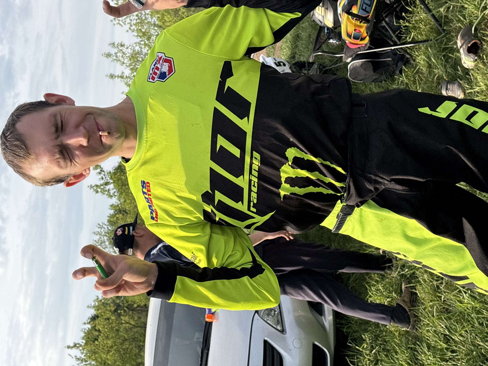 A man is outdoors in a grassy area, wearing a black and fluorescent yellow racing suit with "JORACING" printed on it and Monster Energy branding. He has a cigarette in his mouth and is making a hand gesture. Another person is partially visible behind him near the front of a white van. Racing gear and shoes are scattered on the grass. The location appears to be near a wooded area with a cloudy sky. The location in the city of Votkinsk, Russia cannot be determined based on the visual information provided.
