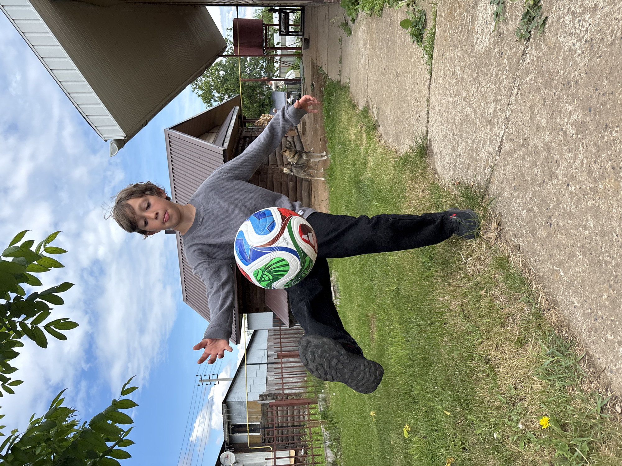 A boy with curly brown hair wearing a gray long-sleeve shirt and black pants is juggling a soccer ball outdoors. He is kicking the ball with his right foot, and his left arm is extended to the side. The soccer ball is brightly colored with blue, green, red, and white panels. The location appears to be a yard next to a concrete walkway and a building with a metal roof. In the background, there is another building with a fence. The sky is visible with blue patches and some clouds. Based on visual details, the general architecture and style of the buildings, the image may have been taken in a rural area of Chaikovsky, Russia. A dog is visible behind the boy.