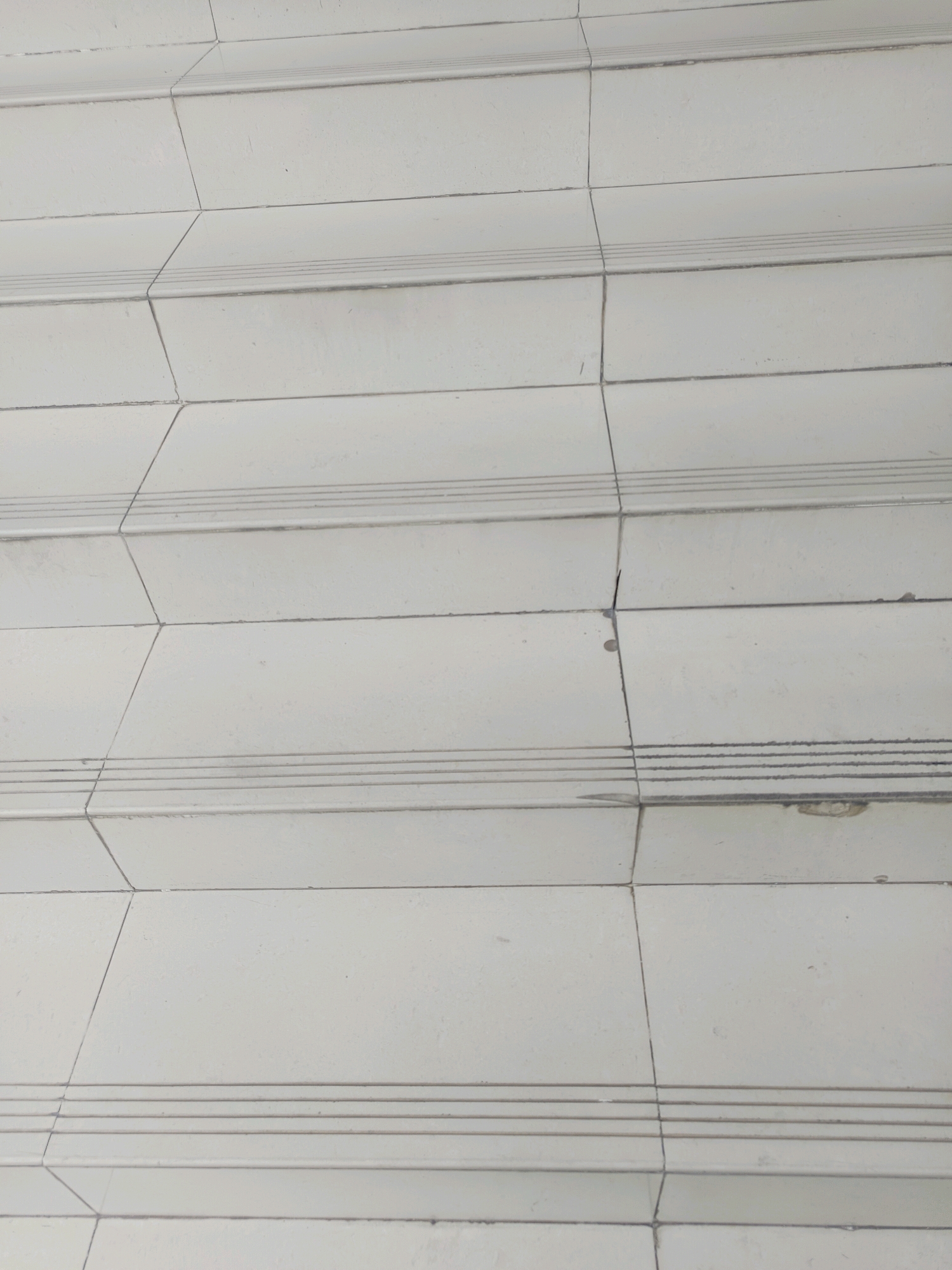 The image shows a close-up view of a tiled staircase. The tiles are white and appear to be worn, with visible grout lines and some minor chips or imperfections. There are horizontal indentations or grooves along the front edge of each step, likely for grip. The staircase is oriented diagonally within the frame, suggesting the viewer is looking down or up at an angle. The overall impression is of an outdoor or indoor public space with functional, utilitarian construction. There are no people or any other identifiable objects or actions in the scene. The location in Sampang, Indonesia, is not discernible from the visual information provided.