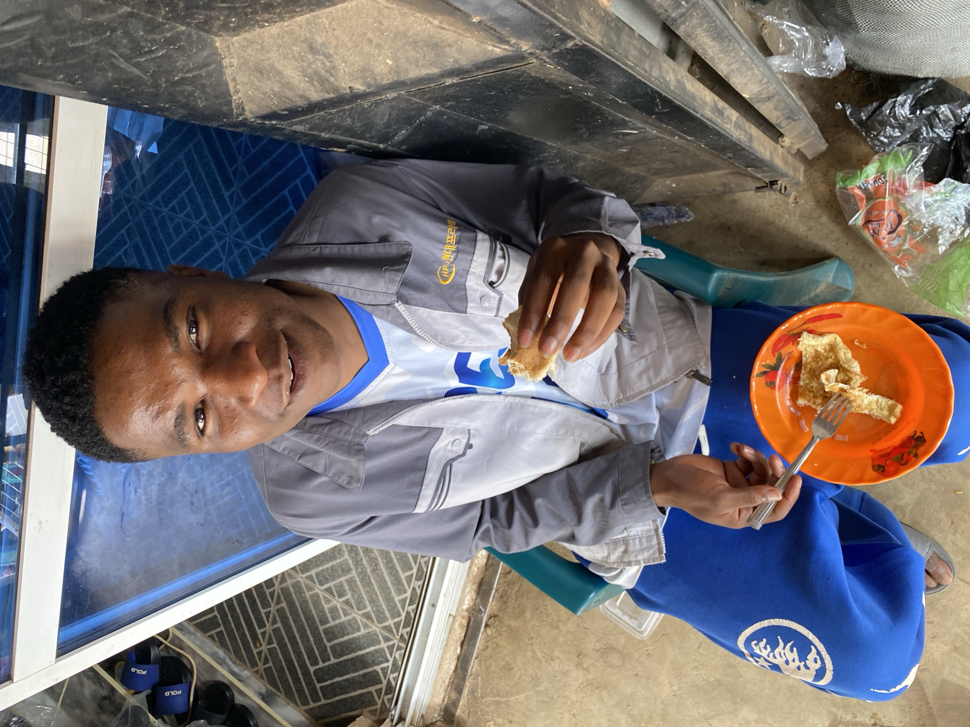 A young Black male is seated outdoors in Bukuru, Nigeria, holding a piece of food and a fork. He is wearing a gray jacket over a blue and white t-shirt and bright blue sweatpants with a white circular emblem on the left leg. A small orange plate with food is in his lap. The background shows a blue tiled wall and a black metal structure. Various plastic bags and footwear are visible on the ground.