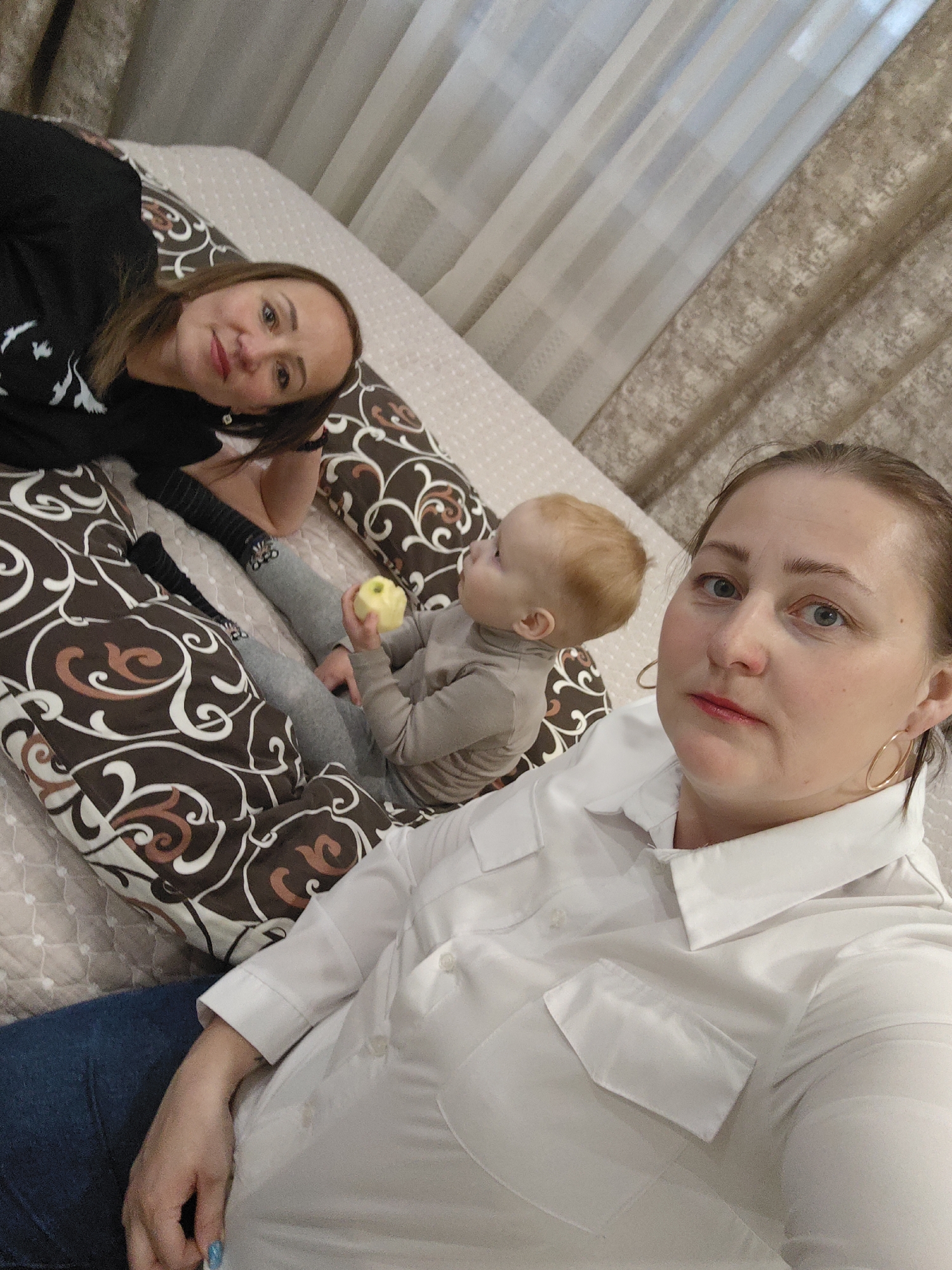 An indoor photograph captures two adult women and a young child relaxing together, likely in a bedroom or living area in Марковский, Russia. In the foreground, a woman with light brown hair, wearing a white collared shirt and blue jeans, takes a selfie or photo. She has a neutral expression, light red lipstick, and golden hoop earrings. Behind her, another woman with dark brown hair lies on a large, U-shaped pillow with a brown, white, and orange patterned cover, wearing a black long-sleeved shirt with white designs. She looks towards the camera with a slight smile. Beside her, a toddler with reddish-blonde hair sits on the same patterned pillow, wearing a grey long-sleeved top and pants. The child holds a green and yellow apple and is looking away from the camera. The individuals are positioned on a light-colored, possibly quilted bed or mattress. The background features light-colored sheer curtains and patterned brown curtains, indicating a comfortable indoor setting. The image suggests a peaceful moment of family or shared relaxation.