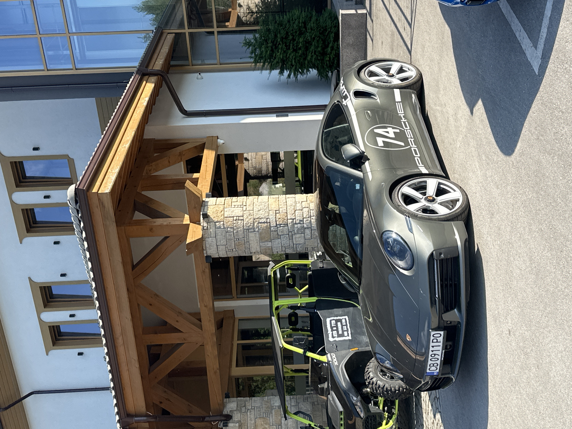 The image shows a grey Porsche 911 Turbo S parked on a paved surface in front of a modern building with a wooden roof structure. The building is constructed with stone pillars and white walls, and features large windows. A small evergreen tree is visible to the right of the building. In the background, a black and neon green ATV is parked. The Porsche has a racing stripe and the number "74" on its side. The license plate of the Porsche is visible and reads "CB 0911 PO". The scene appears to be outdoors during the daytime. The location is the city of Kerenite, Bulgaria.