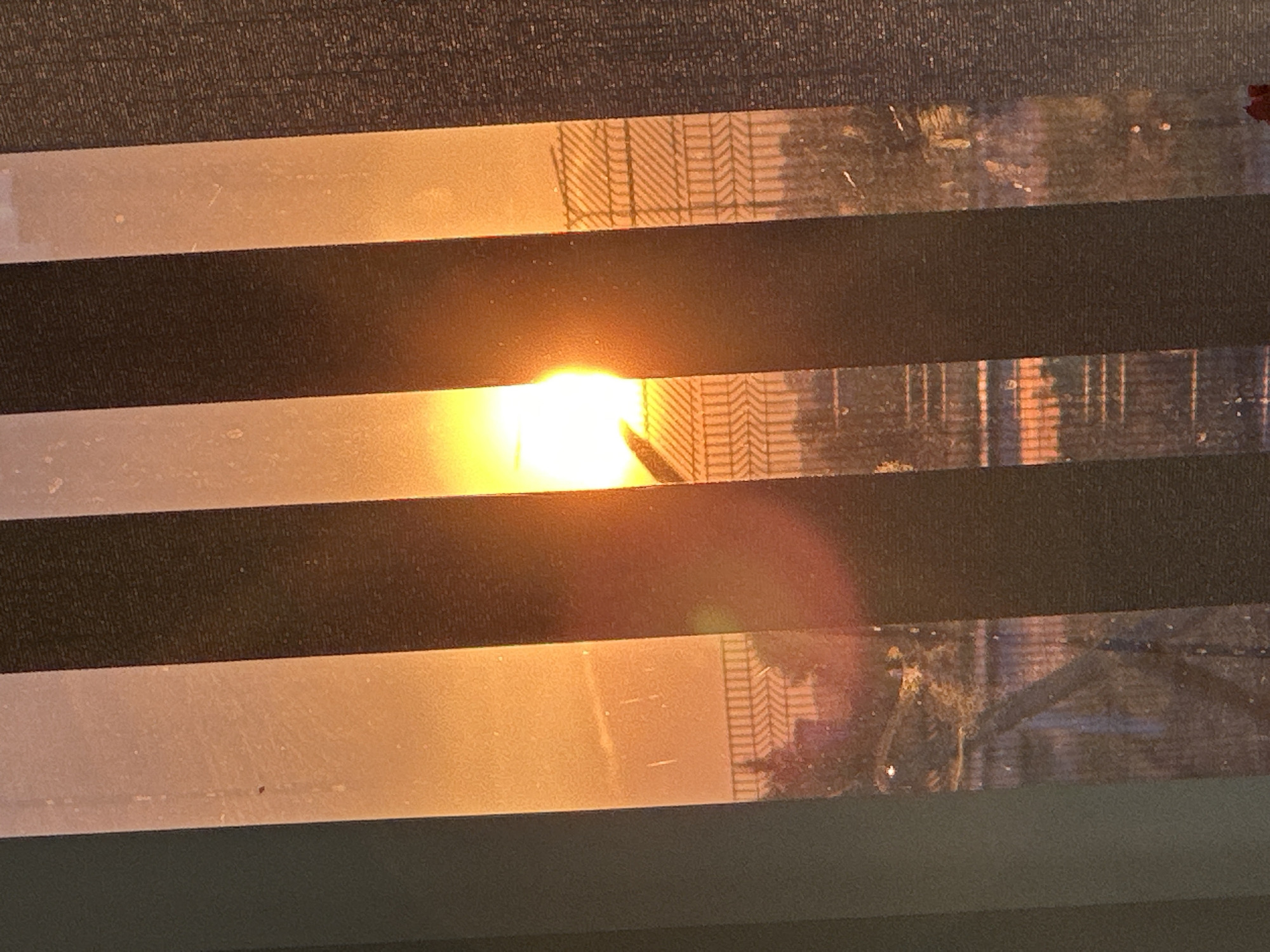 The image depicts a sunset viewed through Venetian blinds. The sun is partially obscured by a dark object, possibly a bird in silhouette, and its rays create an orange glow across the sky. The blinds cast vertical shadows, dividing the scene into alternating light and dark stripes. In the background, a fence with a diamond pattern is visible, along with some foliage and buildings. The overall impression is one of a peaceful evening in an urban setting, possibly in Hora, Ukraine, given the specific contextual information.