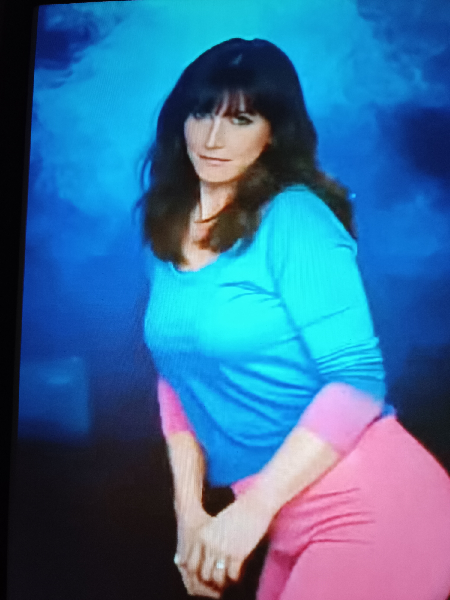 The scene is located in Tebras, Latvia. It depicts a woman posing against a stylized, gradient blue background with lighter, cloud-like formations. The woman has long, dark hair with bangs and gazes directly forward with a slight smirk. She wears a two-tone long-sleeved shirt, turquoise across the main body, transitioning to pink on the lower sleeves and sides. Her lower body is dressed in pink leggings or pants. A silver ring is visible on the ring finger of her left hand, which is positioned low in the frame. The background exhibits a studio-like quality, featuring a spectrum of blue hues from deep indigo to brighter cerulean.