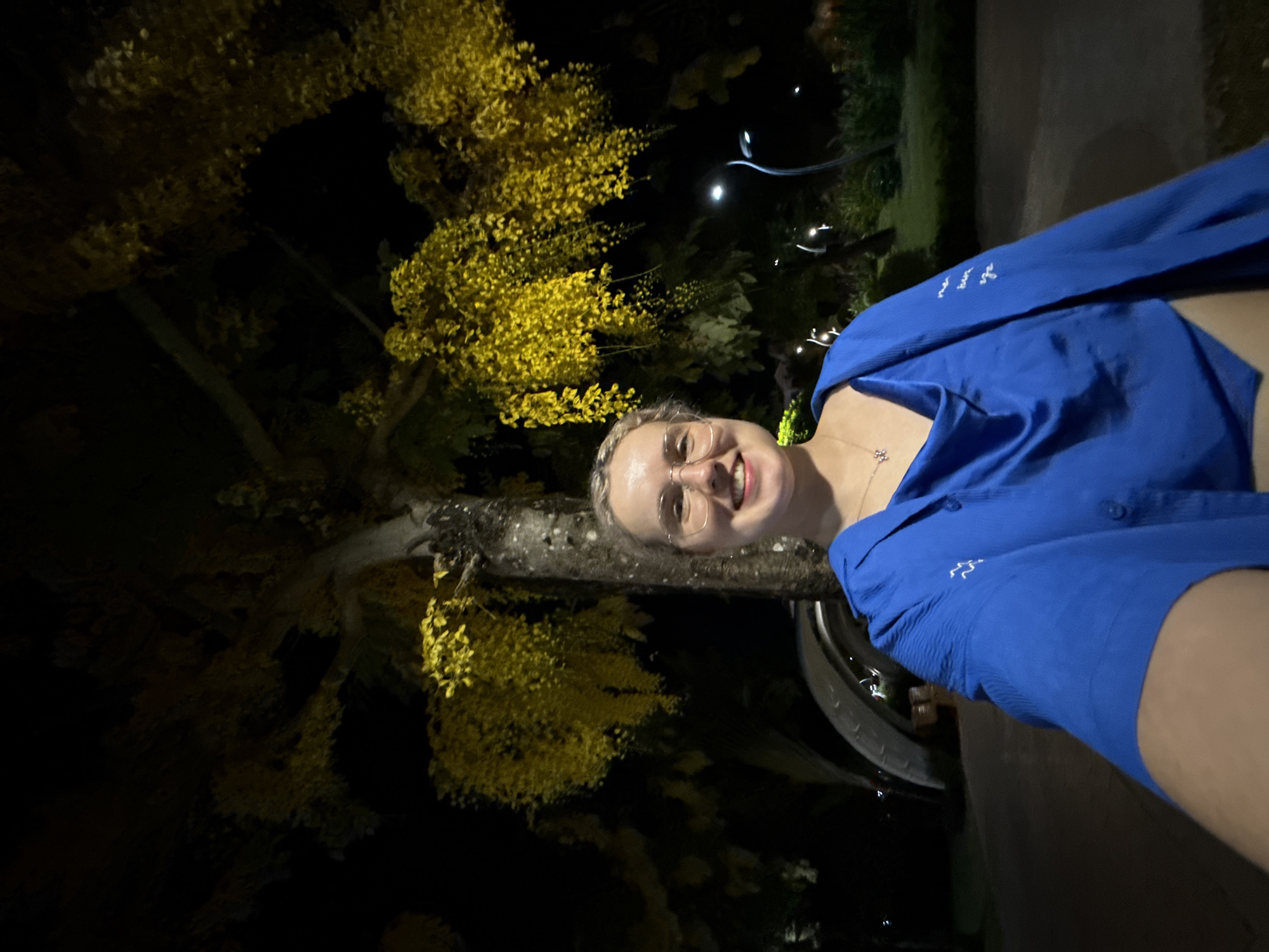 An outdoor, vertical selfie-style photograph taken at night captures a smiling woman against a backdrop of a vividly yellow-flowered tree. The woman, positioned on the right side of the frame, has light-colored hair and wears glasses. She is dressed in a bright blue button-up shirt, which features a small white embroidered Arc'teryx logo on the left sleeve. A delicate silver necklace with a small pendant is visible around her neck.
The dominant feature of the background is a large tree with abundant clusters of yellow leaves or flowers, brightly illuminated against the dark night sky. The tree's trunk is visible, extending diagonally across the frame. In the dimmer, distant background, several small, artificial light sources twinkle, suggesting streetlights or decorative lighting. Part of a dark, circular object resembling a vehicle's tire is partially visible near the bottom right, next to the woman. The overall mood conveyed by the woman's broad smile is cheerful. The setting, characterized by the tropical-looking tree and night lighting, aligns with the provided context of Phú Quốc, Vietnam.