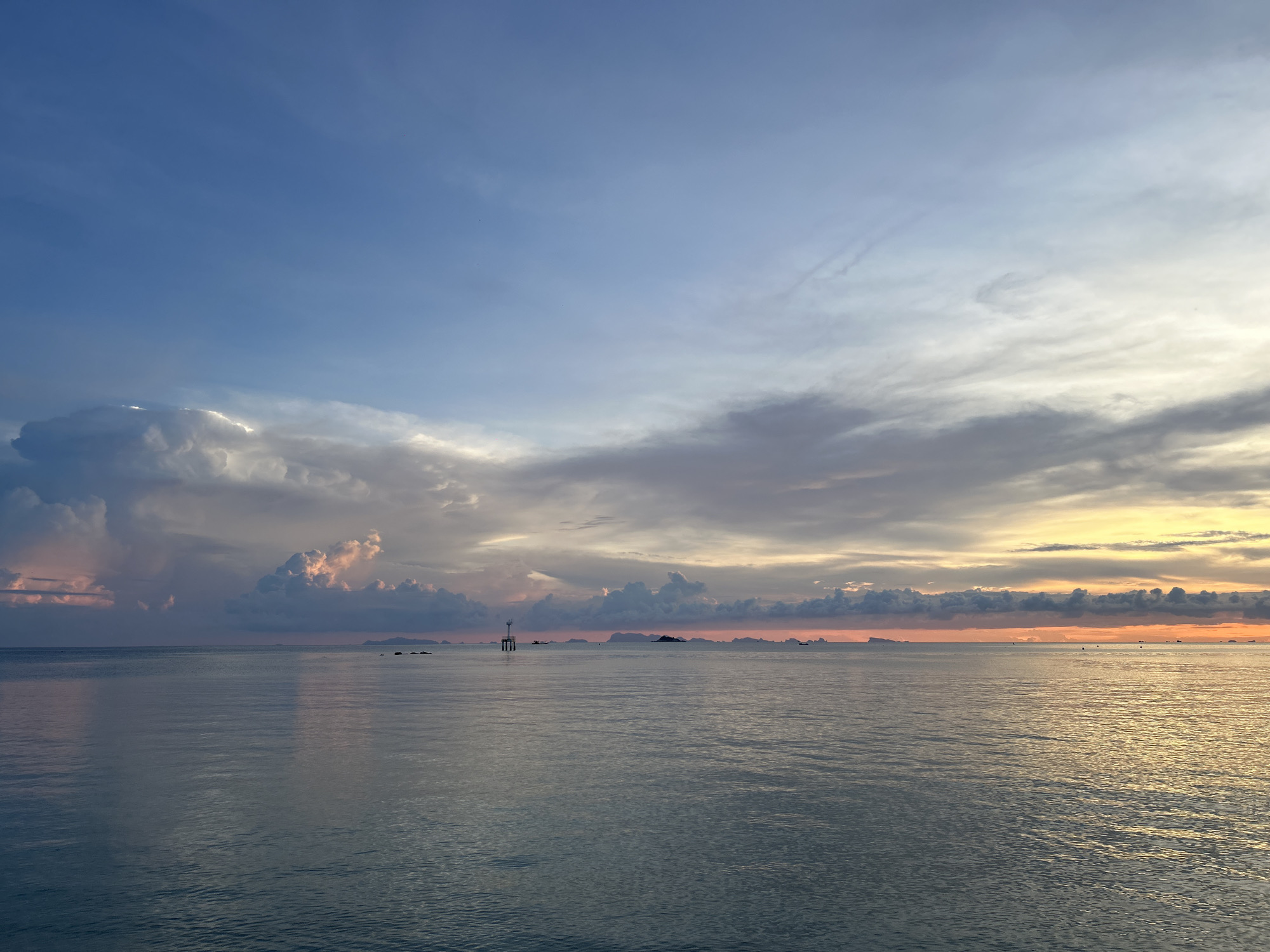 The image depicts a serene seascape at Baan Sri Thanu, Thailand, under a pastel-colored sky transitioning from blue to light orange, indicating either sunrise or sunset. The sea is calm with subtle ripples, reflecting the colors of the sky. In the distance, there are a few small islands or rock formations silhouetted against the horizon, appearing as dark shapes. A white navigational beacon is visible in the water, situated slightly to the left of the center. The clouds are scattered across the sky, with some areas showing a pinkish hue from the light. There are no people visible in the image.