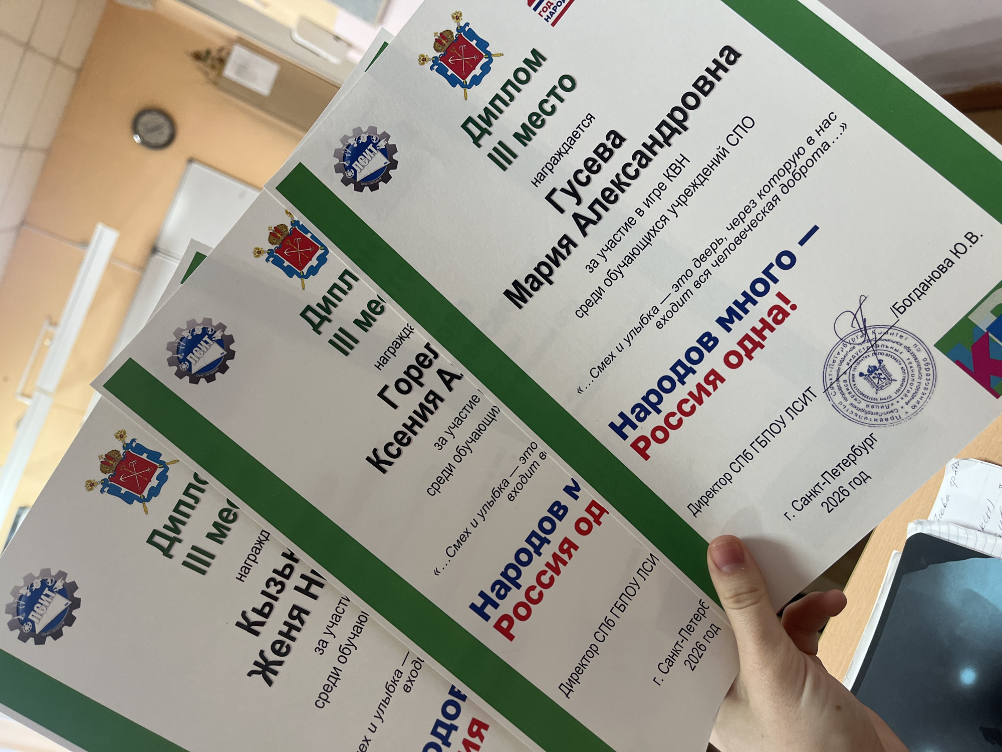 Here's a detailed description of the image:
The image displays three certificates ("Диплом III место" - Diploma III Place) held in a person's hand. The certificates are for third place winners in a competition related to the game KVN (a comedy competition), amongst students of secondary vocational education institutions (СПО). The recipients are:
1. Maria Alexandrovna Guseva
2. Ksenia A. Gorel
3. Zhenya N. Kyzn
All certificates include the phrase "Народов много - Россия одна!" (Many peoples - Russia is one!). They are signed by the director of СПб ГБПОУ ЛСИТ (Saint Petersburg State Budgetary Professional Educational Institution, Lyceum of Service and Industrial Technologies). The certificates state that they were issued in Saint Petersburg in 2026. A portion of a notebook and a tablet are seen in the background.