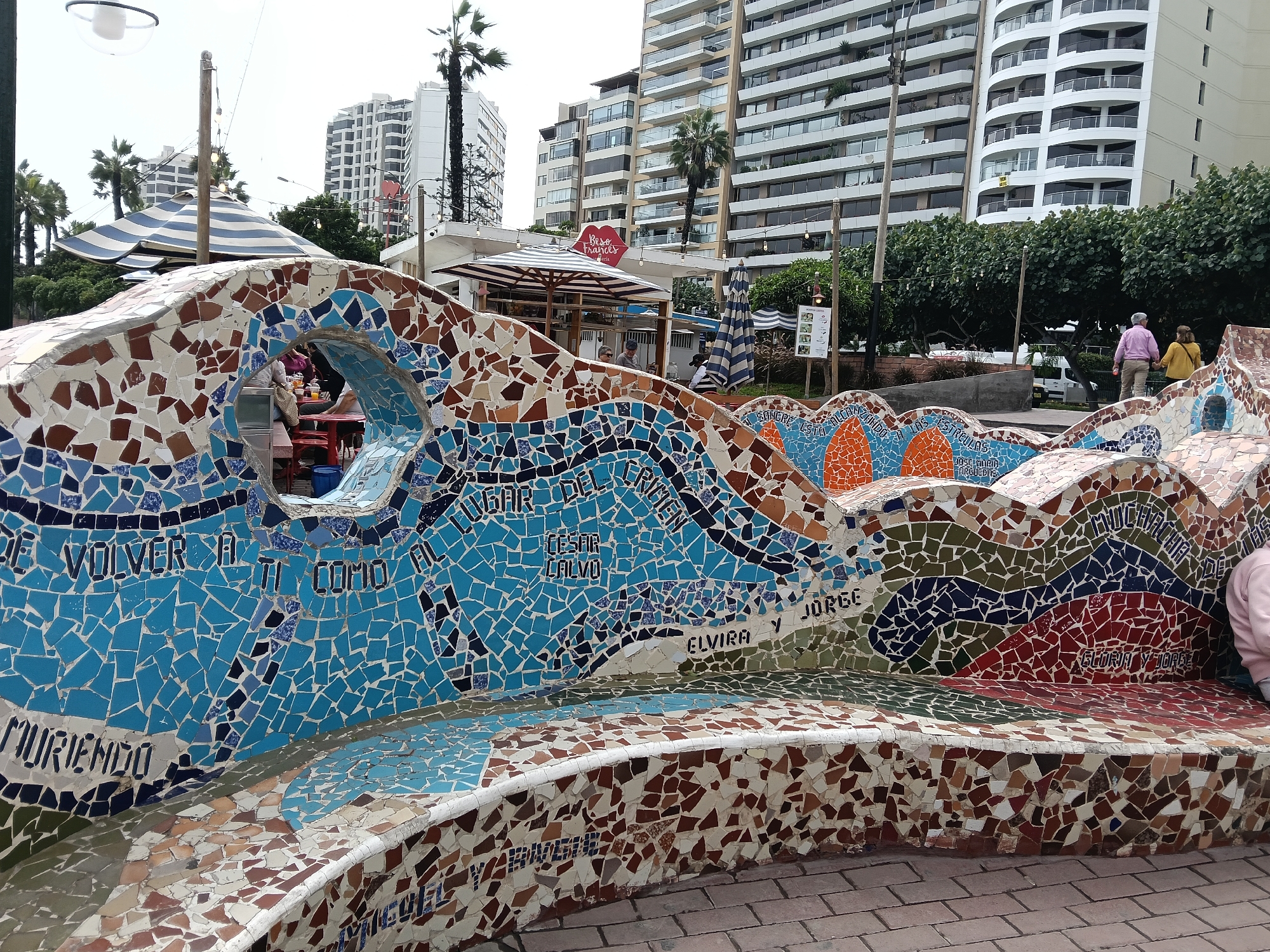 Here is a description of the image:
The image captures a vibrant, mosaic-covered bench in a public space, likely a park, in Miraflores, Peru. The bench is adorned with colorful tiles, primarily in shades of blue, brown, green, orange and red. Inscribed within the mosaic are phrases and names like "De volver a ti como al lugar del crimen," "Cesar Calvo," "Elvira y Jorge," "Muriendo," "Gloria y Jorge," and "Beso Frances." The bench curves in a serpentine manner.
In the background, there are several multi-story buildings, lined with balconies, and palm trees. There is a kiosk-like structure with striped awnings, and a red sign, suggesting a food or beverage stand. Two people are walking in the background. The overall setting suggests a sunny day, and a relaxed, public environment. The scene is characterized by a mix of art, public amenity, and urban landscape.