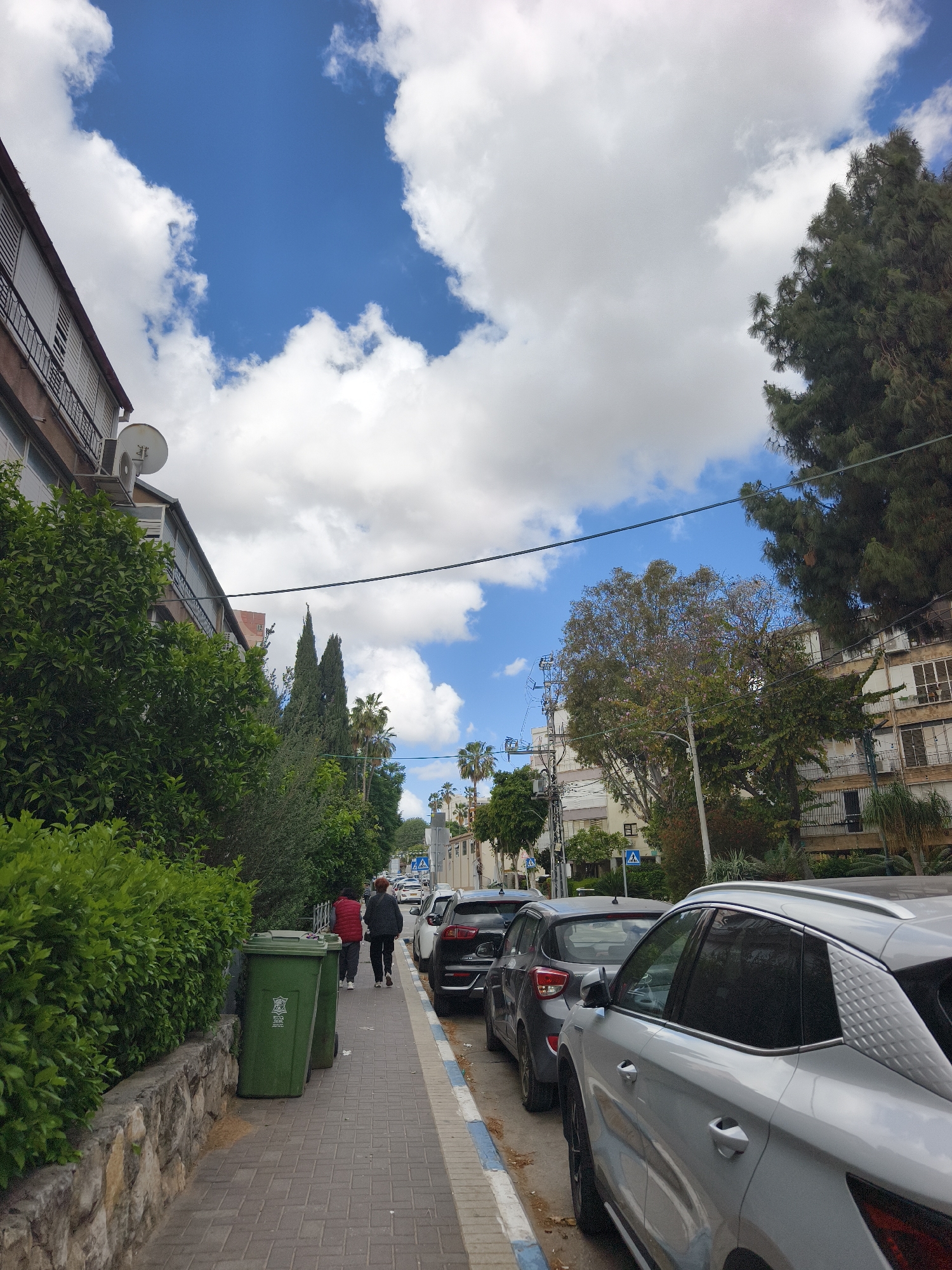 A sunny day in Petah Tikva, Israel. A tree-lined residential street features a paved sidewalk, parked cars, and buildings on either side. Two people walk along the sidewalk near green trash bins. The sky is bright blue with scattered white clouds. Power lines stretch across the sky. Buildings are visible on both sides of the street, and the parked cars line the curb.