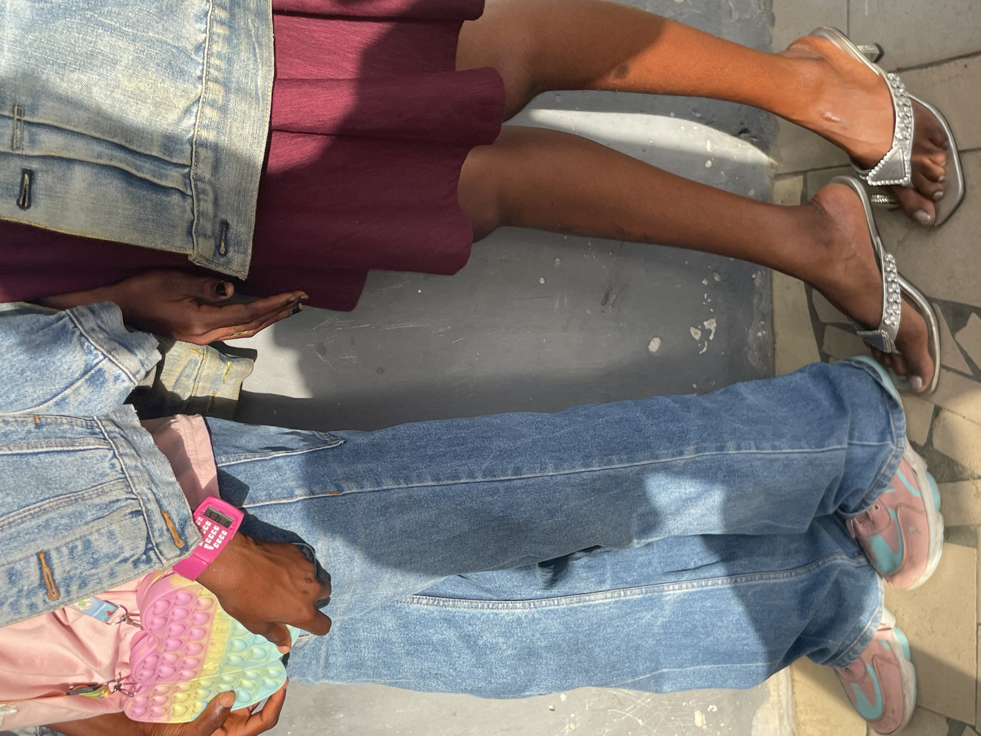 Two individuals are shown from an overhead perspective, lying or sitting on a grey, textured ground in Badagry, Nigeria, under bright daylight conditions.
The person in the upper half of the frame wears a light blue denim jacket over a maroon pleated skirt. Their hand, adorned with dark nail polish and a subtle gold ring or bracelet, rests on the ground. Their legs and feet, displaying a dark skin tone, are clad in silver high-heeled sandals embellished with rhinestones.
The person in the lower half wears a light blue denim jacket over light blue, baggy jeans, with pink and light blue sneakers on their feet. Their hand, also of a dark skin tone, is holding a heart-shaped pop-it fidget toy in shades of pink, yellow, and light blue. A pink calculator watch, displaying the text "SMS" on its screen, is visible on their wrist.
Both individuals appear to be in a relaxed, casual posture, suggesting an activity of leisure or rest.