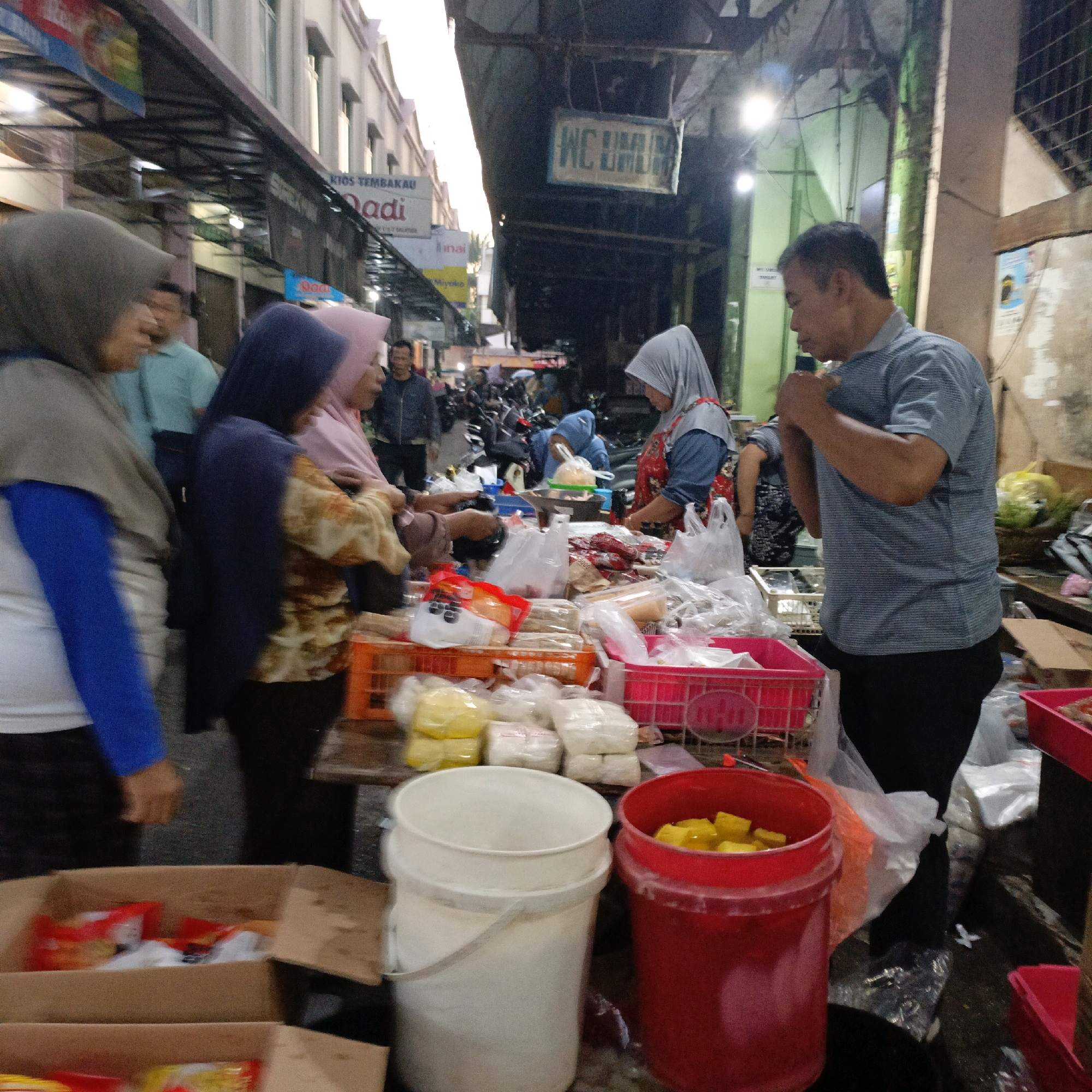 The image depicts a bustling street market scene, likely in Salatiga, Indonesia, given the Indonesian text visible on signs. The time appears to be late afternoon or early evening, judging by the artificial lighting and the overall dimness. In the foreground, a variety of market stalls are laden with goods. Several individuals are engaged in commerce. On the left, a woman wearing a grey hijab and a blue long-sleeved shirt is interacting with the market. To her right, another woman in a dark blue hijab and a patterned shirt is also present. Further right, a woman in a light pink hijab and an apron is attending to her stall, which is filled with packaged goods. A man in a striped grey shirt is also a prominent figure, looking towards the left and gesturing with his hands.
The stalls display a range of items, including what appears to be food products in plastic packaging, some in yellow and white. There are also red and white buckets filled with yellow cubes, possibly food items. Cardboard boxes containing packaged goods are also visible. In the background, more market activity is implied, with the presence of motorcycles, suggesting a busy thoroughfare. Signs with Indonesian text, such as "WC UMUM" (Public Toilet) and "Kios Tembakau" (Tobacco Kiosk), provide context about the commercial nature of the location. The environment seems to be an open-air market with a covered section, characterized by makeshift structures and a somewhat cluttered ground, hinting at the daily operations of local vendors and shoppers.