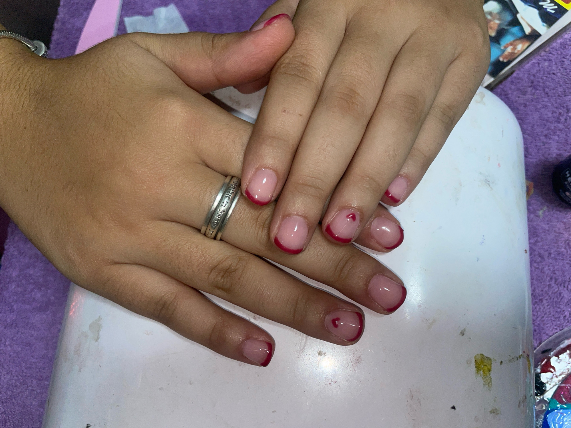 The image shows a close-up of a person's hands with a manicure. The nails are short and square-shaped, with a clear base coat and a French tip in a dark pink or red color. Some of the nails also have a small heart shape painted on them in the same dark pink/red color. The person is wearing a silver ring on their left ring finger. The hands are resting on a white, somewhat dirty surface, possibly a nail lamp or a table at a nail salon. In the background, there is a purple towel and some objects that appear to be nail polish bottles and other manicure tools. The lighting suggests an indoor environment. There is no visible indication of the location being in Raanana, Israel.