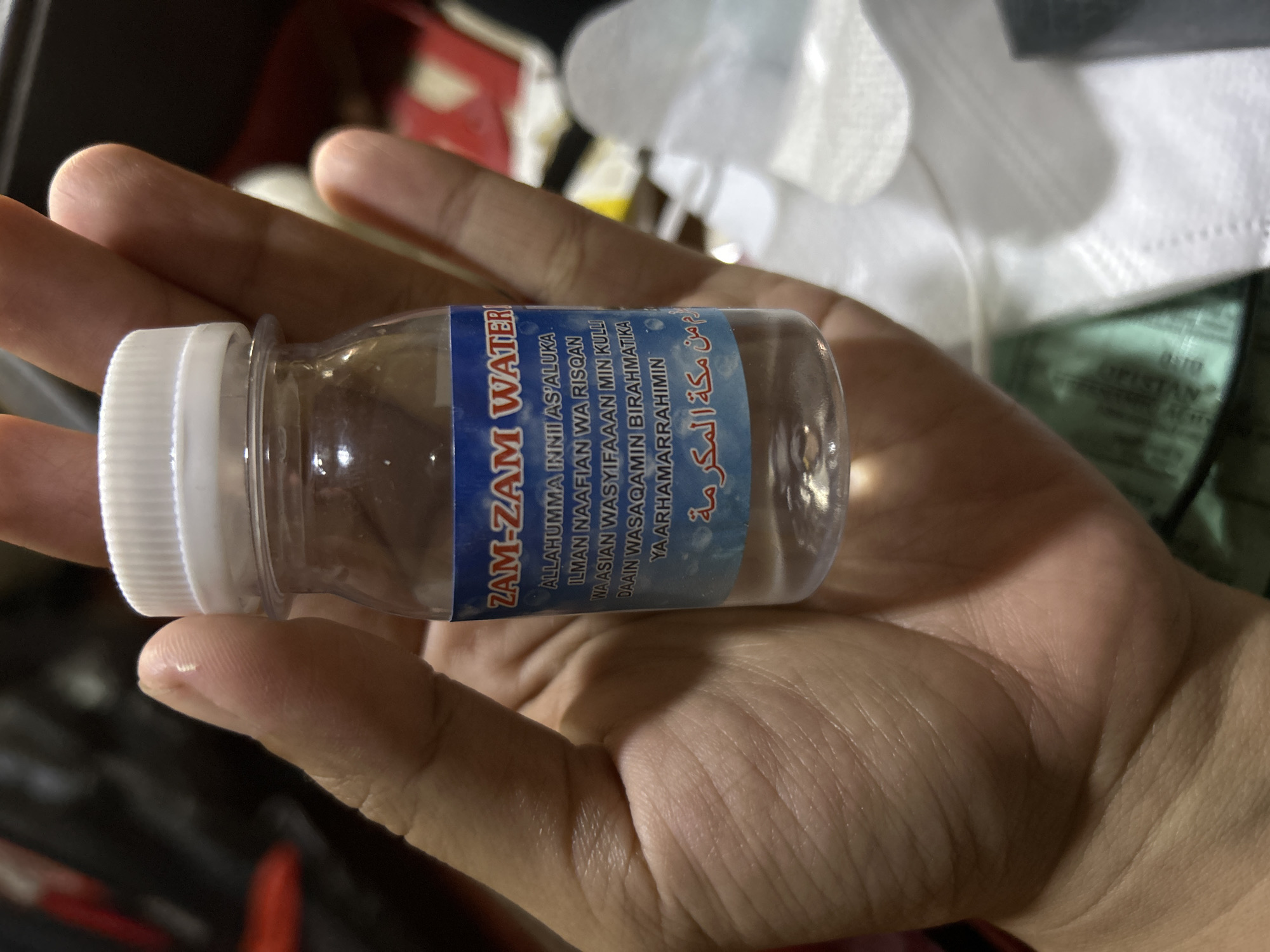 A person's hand is holding a small, clear plastic bottle filled with clear liquid. The bottle has a white cap and a blue label with white text. The text on the label appears to be in both English and Arabic. The English text reads "ZAM-ZAM WATER" followed by a prayer. The Arabic text is below the English text. The hand is a light to medium skin tone and shows finger lines and palm creases. The background is out of focus but appears to be indoors with some dark and lighter colored objects, possibly suggesting a home or market environment. The scene is well-lit, focusing on the bottle and the hand holding it. There is no indication of the specific location within Depok, Indonesia, in the provided image.