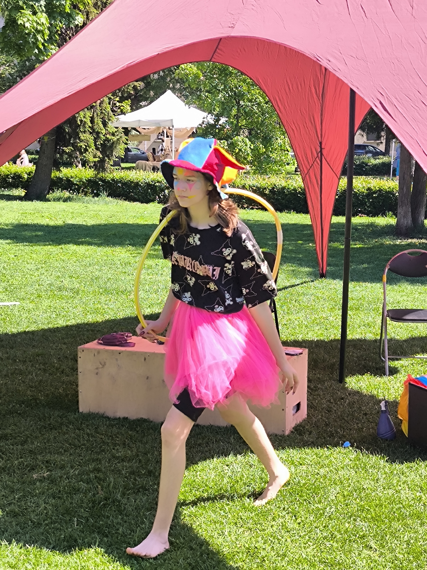 A young barefoot person with face paint walks on grass in a park. The person wears a black t-shirt, a pink tutu, a jester hat, and black shorts. A yellow hula hoop is in their hand. A red tent is overhead. A wooden box is behind the person, and a tent is in the background. The photograph appears to have been taken in Riga, Latvia.