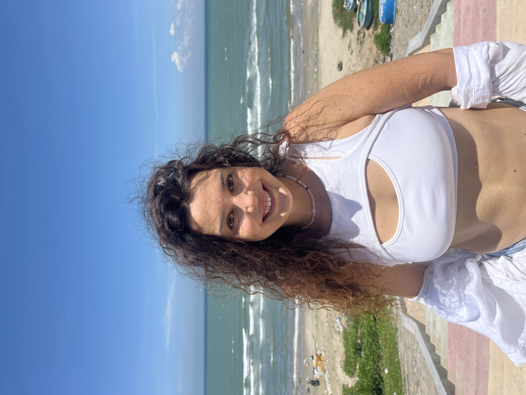 A woman stands on a set of colorful stairs facing the sea. She has dark, curly hair and is smiling at the camera. She is wearing a white crop top with a cut-out detail and a white open shirt draped over her shoulders. The background shows a sandy beach littered with debris, and the ocean meeting a clear blue sky. The location is identified as Mũi Né, Vietnam.