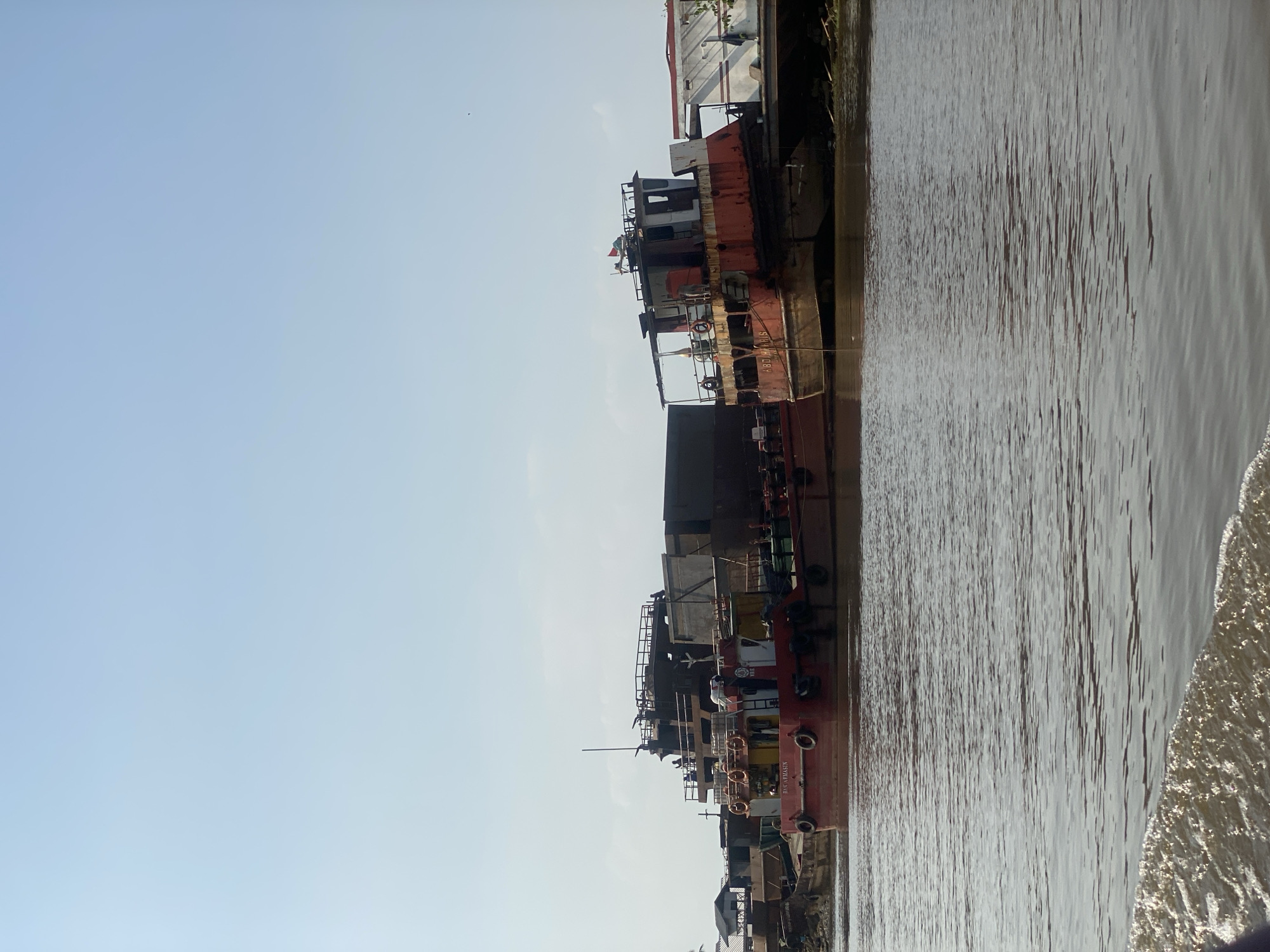 In Handil Bakti, Indonesia, a waterway scene unfolds under a clear, pale blue sky with a few wispy clouds. Several docked barges and boats occupy the muddy brown water, suggesting a port or industrial area. The primary focus is on two large, aged vessels.
The boat on the left is a long, red barge with "BAS ARMASIN" written on its side. It is laden with dark cargo containers. A smaller, red and white boat is positioned in front of the barge. The second, larger vessel to the right, also appears aged and weathered, painted predominantly in faded red and white. It has a prominent superstructure with windows and a covered walkway. A small flag is visible on its roof. Both vessels are moored side-by-side, their hulls resting in the water.
There are several figures visible on the decks of the boats, appearing to be engaged in activities related to the vessels, though their specific actions are not discernible. In the background, a small, simple building with a dark roof is visible on the shore, partially obscured by the boats. The water in the foreground displays ripples and some foaming, indicating the presence of a boat or movement. The overall impression is one of a working waterway, possibly a cargo loading or unloading area.