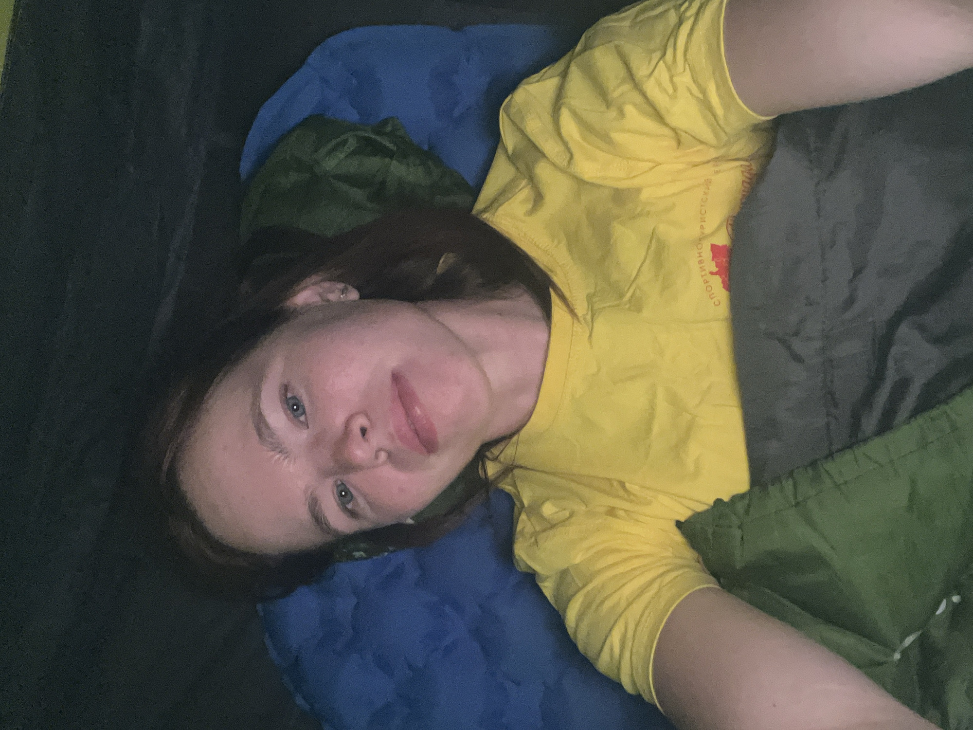 A woman is lying in a sleeping bag inside a tent. The woman has fair skin, light blue eyes, and brown hair. She is wearing a yellow t-shirt with Russian text. The sleeping bag is green and gray. A blue pillow is beneath her head. The interior of the tent is dark. The image was taken indoors. There is no information in the image to confirm the location is Khvalynsk, Russia.