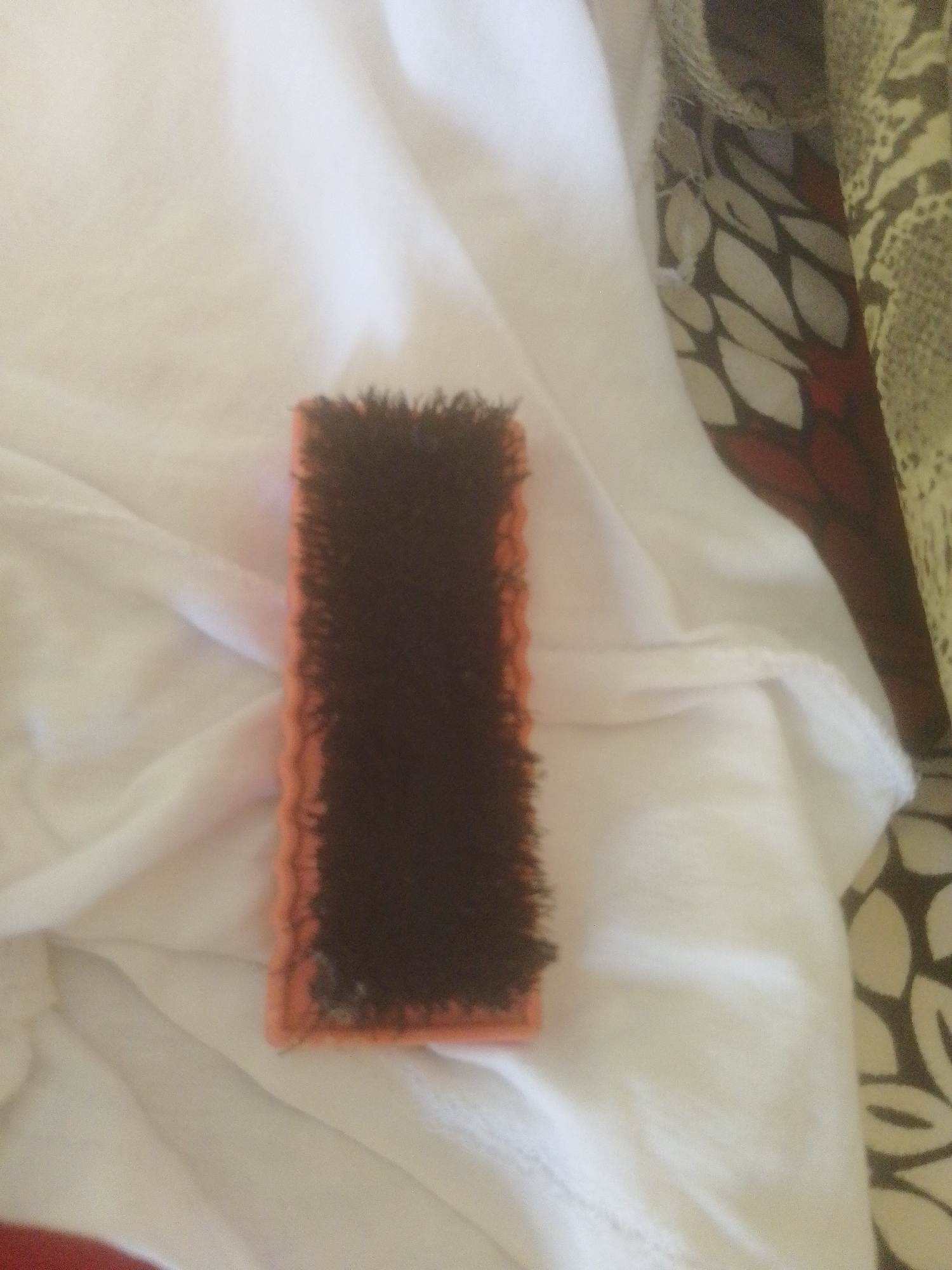 A close-up, slightly out-of-focus, overhead shot captures a bristly, rectangular cleaning brush with an orange plastic base and dark brown bristles. The brush is positioned on top of a white, wrinkled fabric, likely clothing or bedding. To the right of the brush, a patterned fabric with a cream, black, and red design is visible, suggesting it is part of the same item or a nearby garment. The scene is dimly lit, with shadows cast on the white fabric, creating a soft, diffused illumination. The overall impression is that of a domestic setting, possibly a bedroom, where laundry or cleaning supplies are present. There are no people or any discernible city elements visible, making it impossible to ascertain its location in Danbazau, Nigeria, or any other specific geographical area.