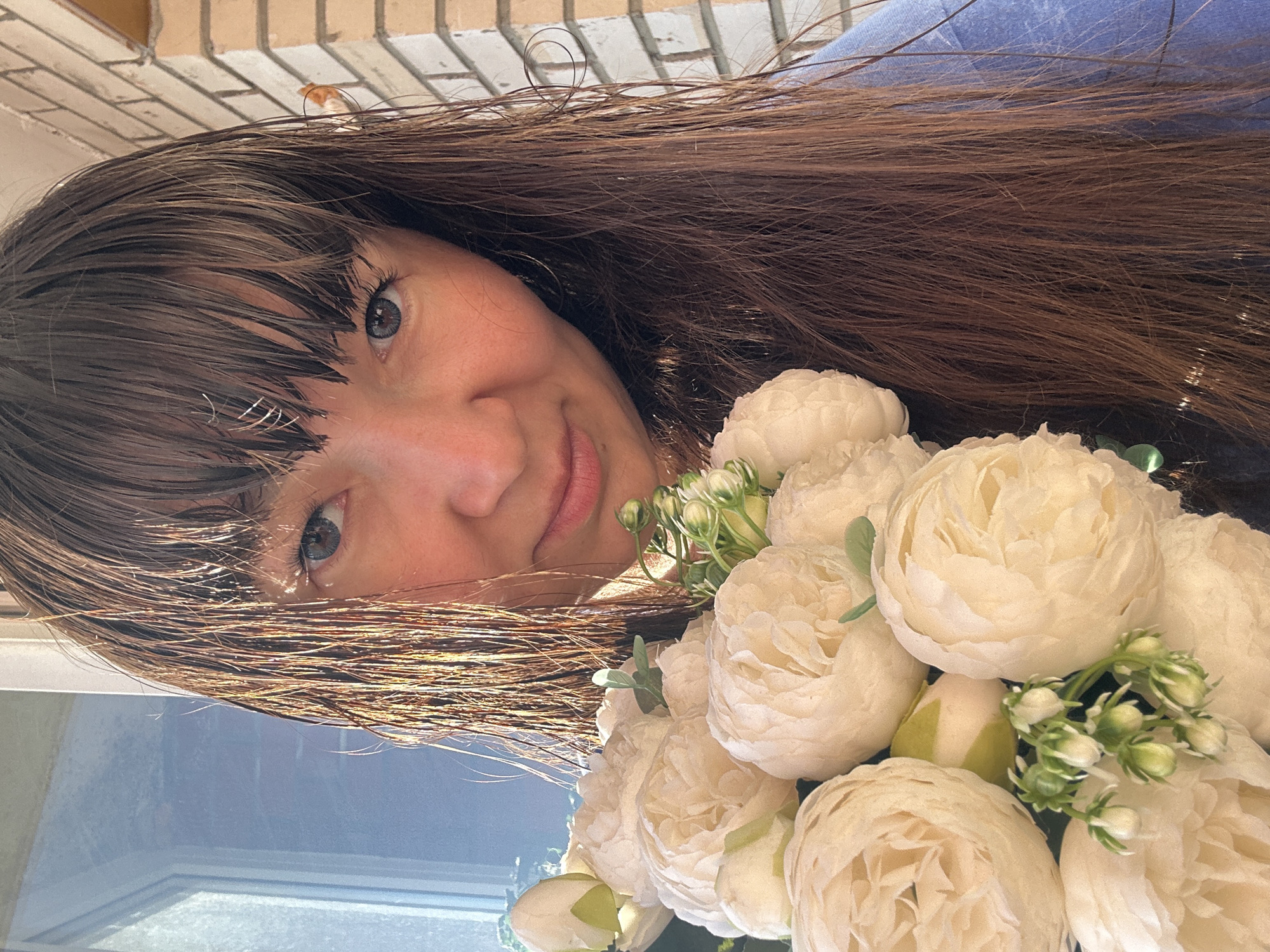 In this close-up, eye-level shot taken outdoors, a woman with long, dark brown hair and bangs is partially obscured by a bouquet of white peonies. She is looking at the camera with a slight smile, her blue eyes visible. The background consists of a white brick wall on the right and a window with vertical blinds on the left, suggesting an urban setting. The natural light illuminates the scene, casting shadows and highlighting strands of hair. The woman is wearing a blue garment. The composition focuses on her face and the flowers, with the surroundings serving as a muted backdrop.