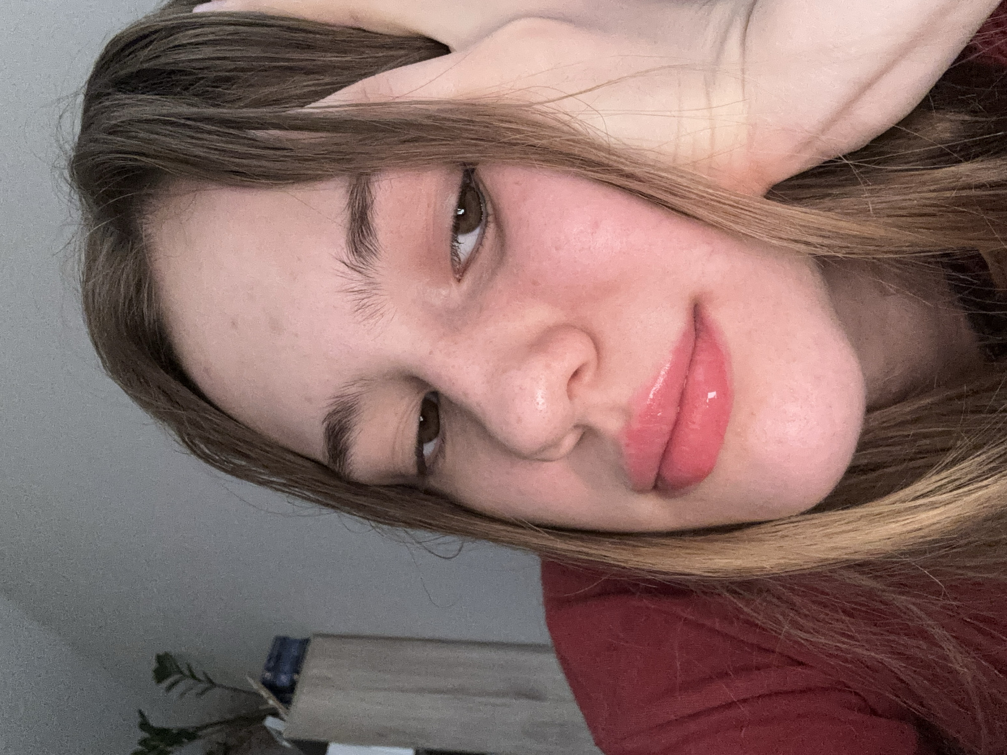 A young woman with light brown hair and freckles rests her head on her hand, her eyes looking downwards. She is wearing a red top. Her lips are glossy and pink. The background is a soft grey wall with a potted plant and some shelving to the left of the frame. The lighting is dim, suggesting an indoor setting.